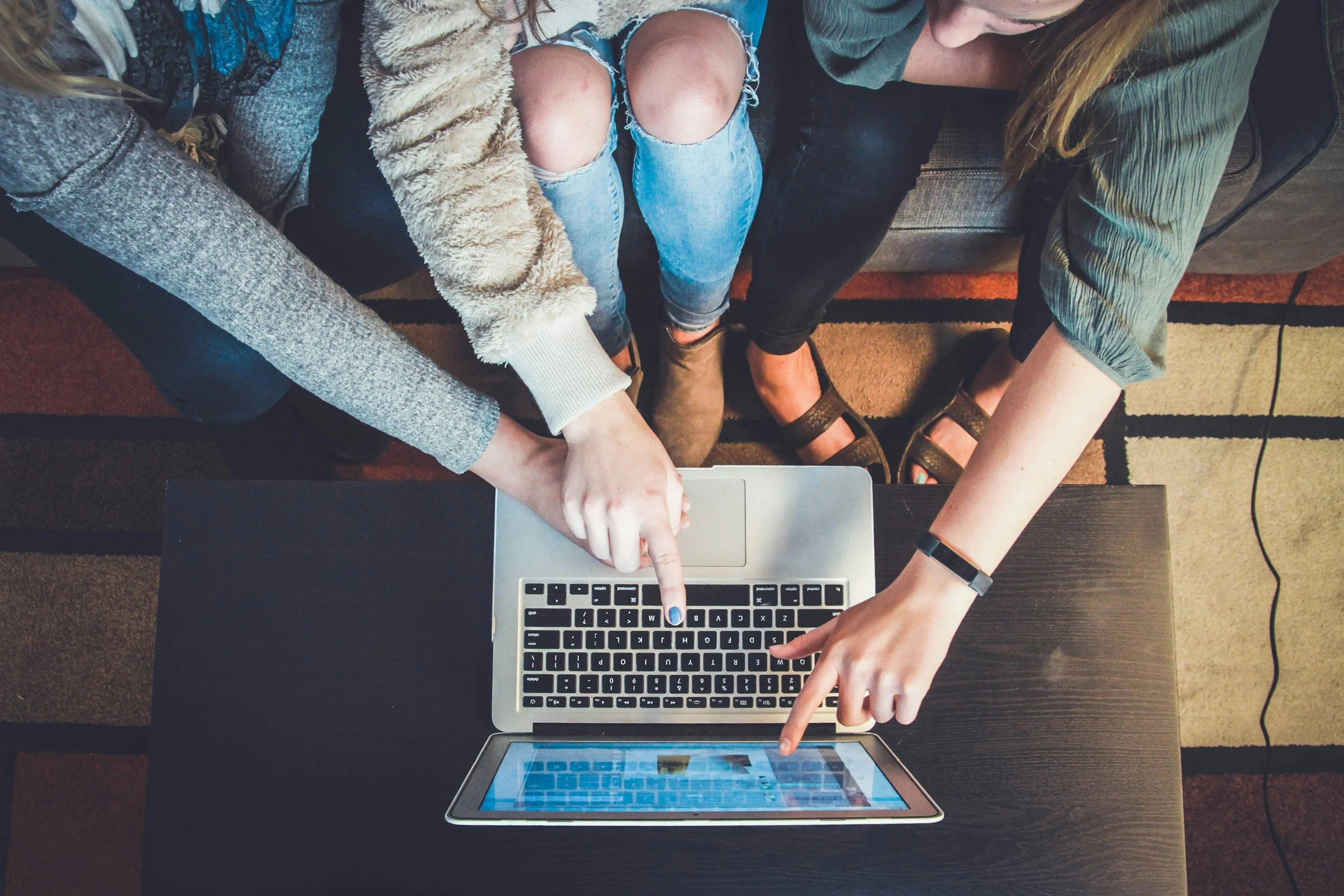 Three people sitting on a couch pointing at a laptop and a tablet on a table.
