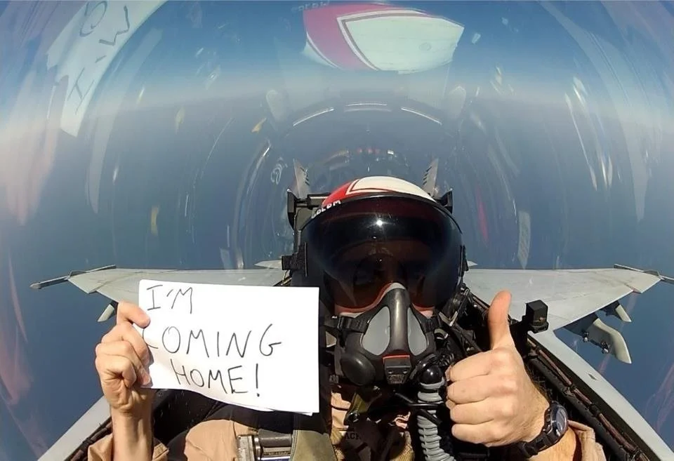 Pilot in fighter jet cockpit holding a sign that says 'I'M COMING HOME!' and giving a thumbs-up.