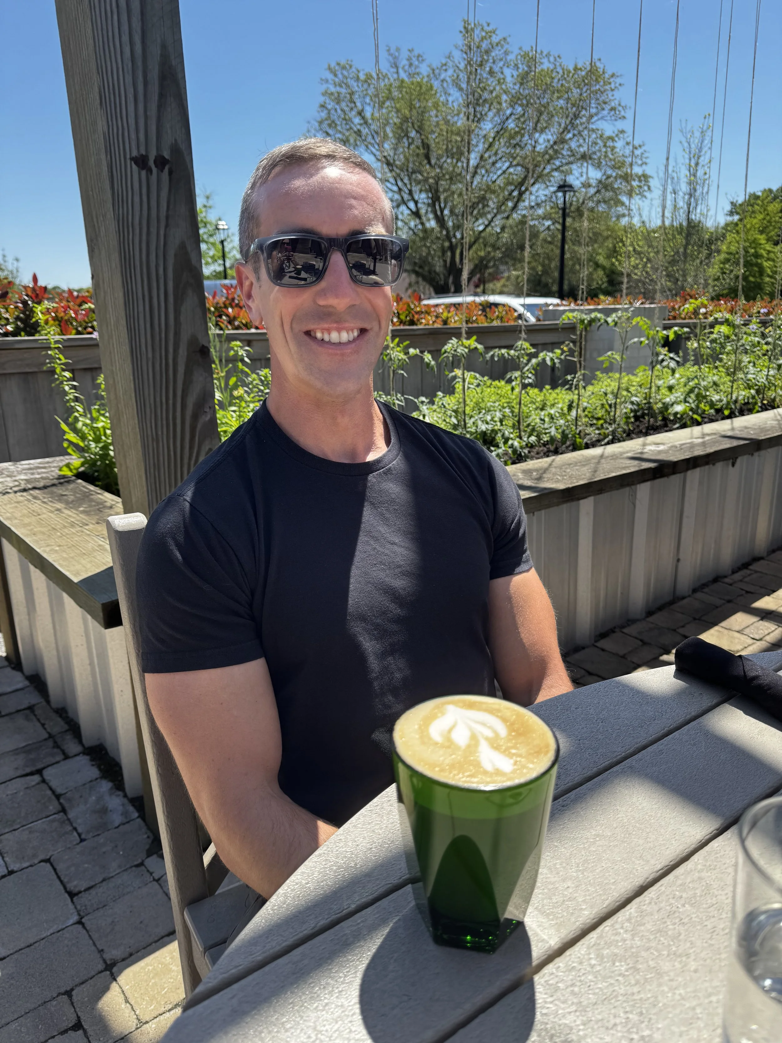 A smiling man wearing sunglasses and a black t-shirt sitting outdoors at a table with a green drink topped with foam and latte art on a sunny day.