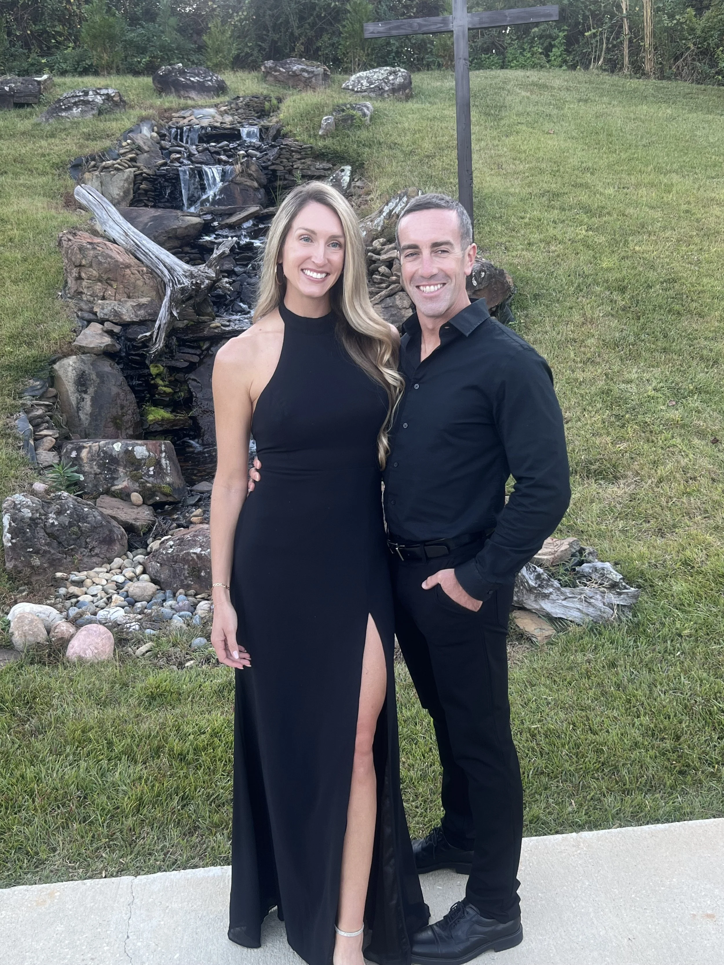 A smiling woman in a black evening gown with a thigh-high slit and a man in a black shirt and pants, standing outdoors on a concrete path in front of a small waterfall and a grassy area with trees.