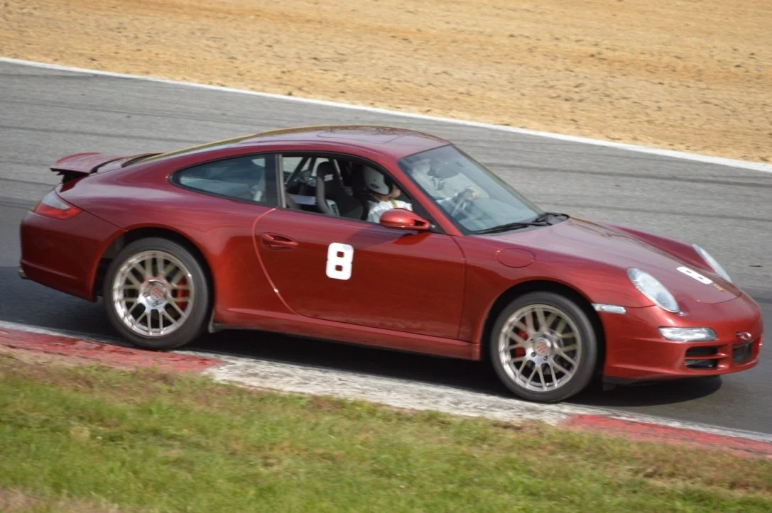 Red sports car on a racetrack with the number 8 on its hood and side, driver wearing a helmet, leaning into a turn.