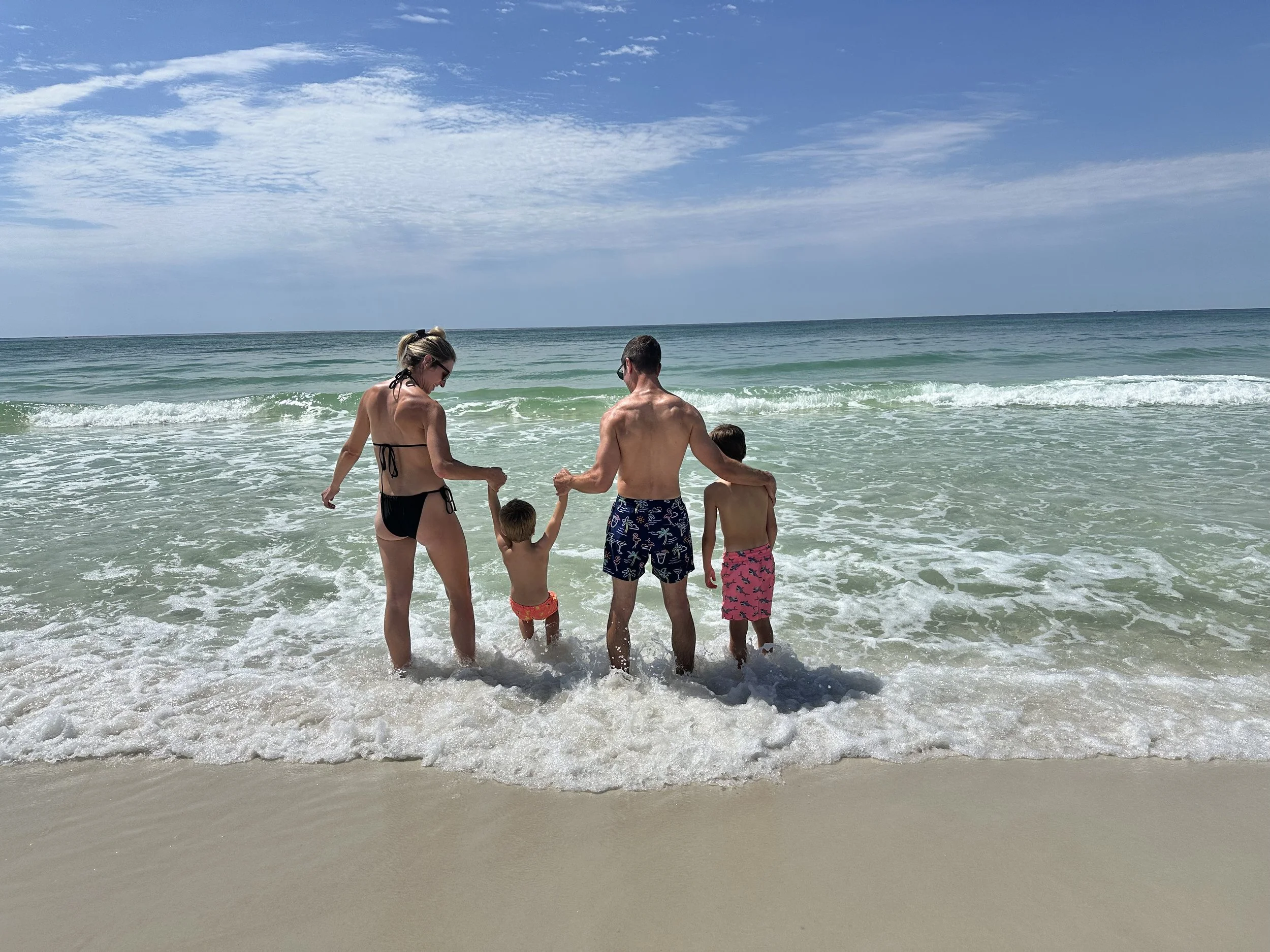 Family of four standing in the ocean waves at the beach, holding hands and facing the water on a sunny day.