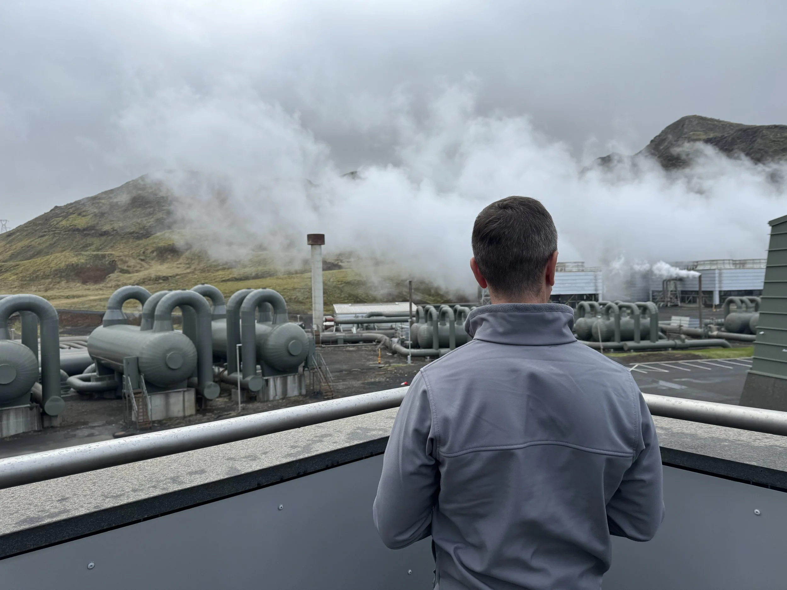 A man wearing a gray jacket stands with his back to the camera, overlooking an industrial site with pipelines and steam emissions against a mountain and cloudy sky.
