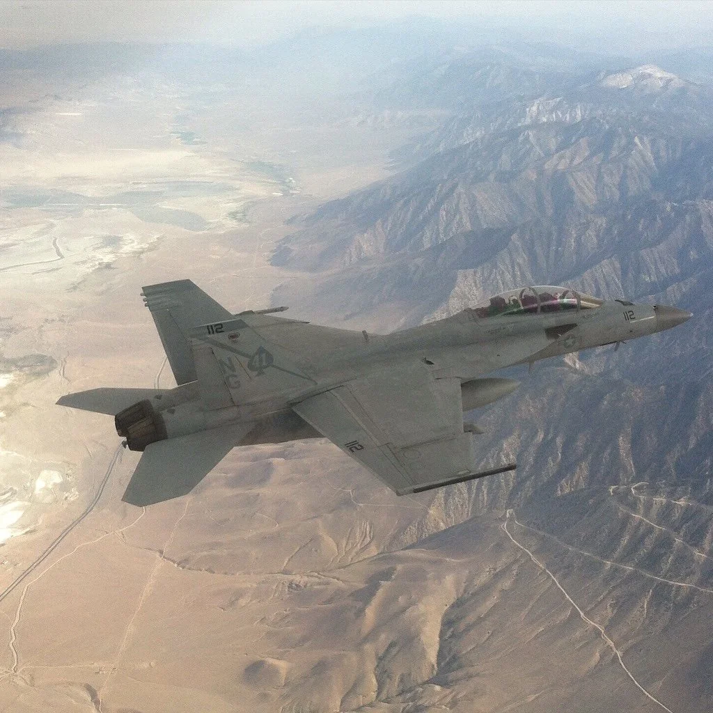 A military fighter jet flying over a mountainous and desert landscape.