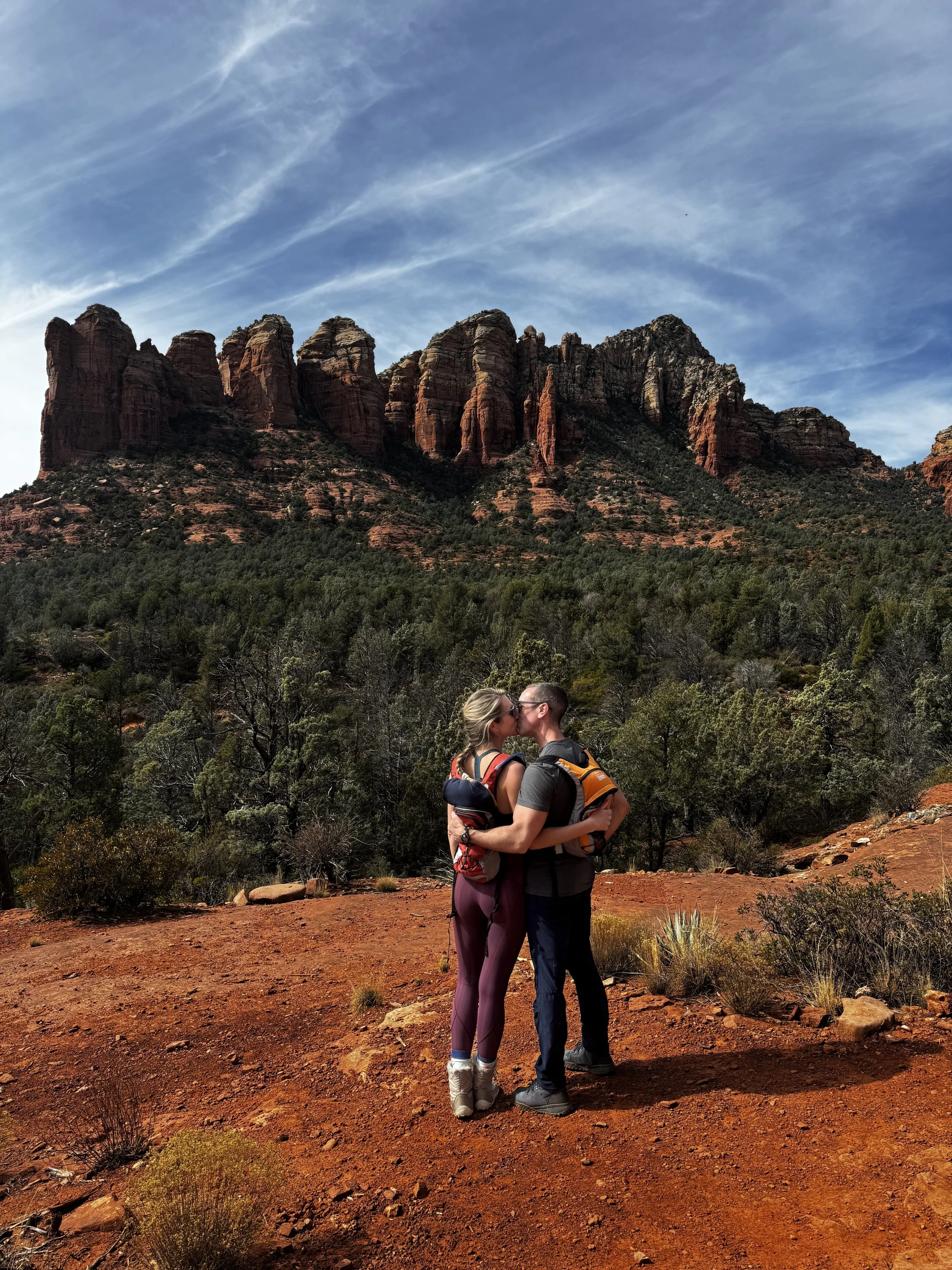 A couple kissing outdoors with mountains and desert vegetation in the background.