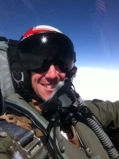 A person in military or aviation gear smiling, wearing a helmet and sunglasses, with a sky and clouds in the background.