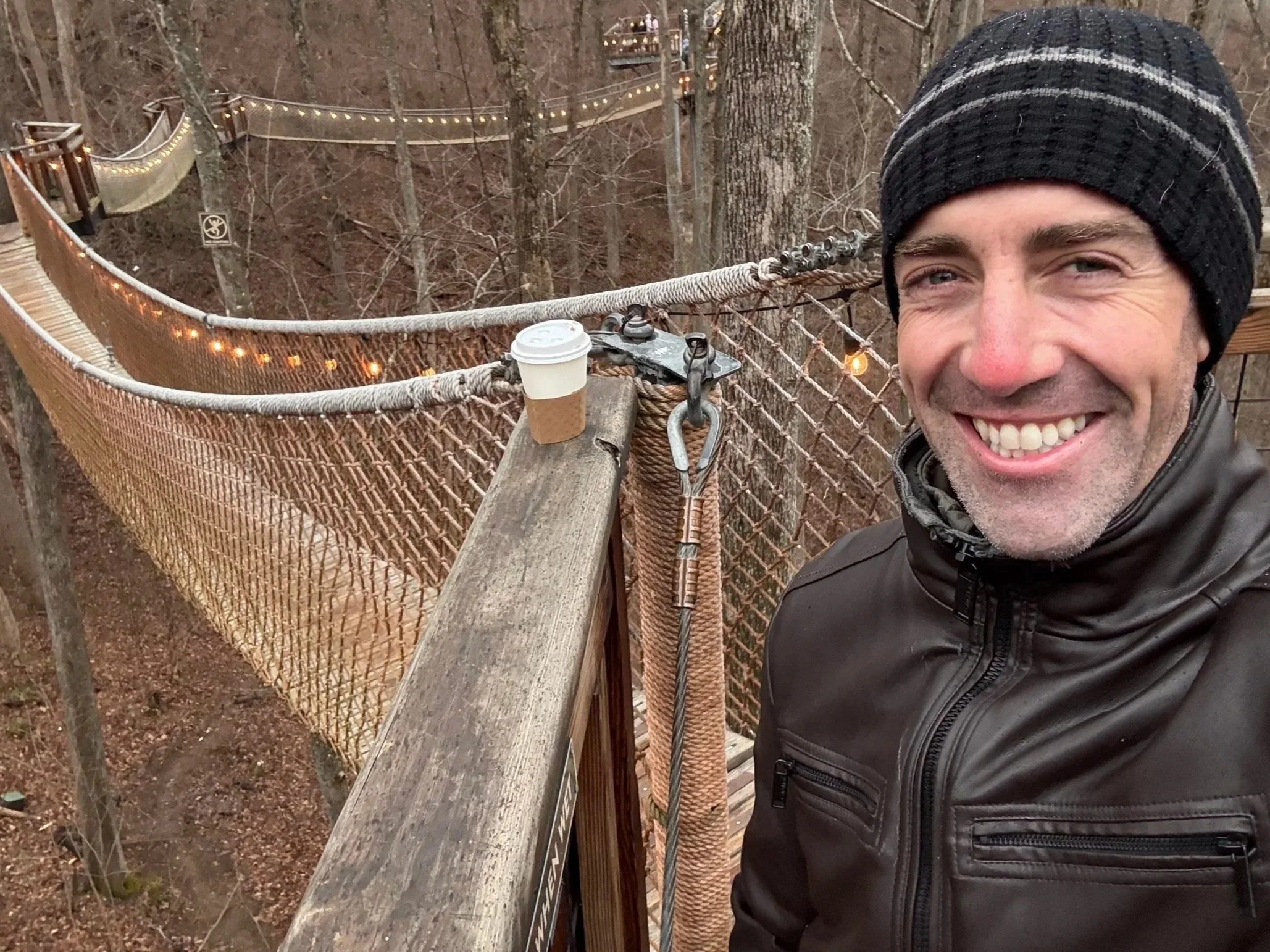 A smiling man taking a selfie on a wooden suspension bridge in a forest with leafless trees and string lights.