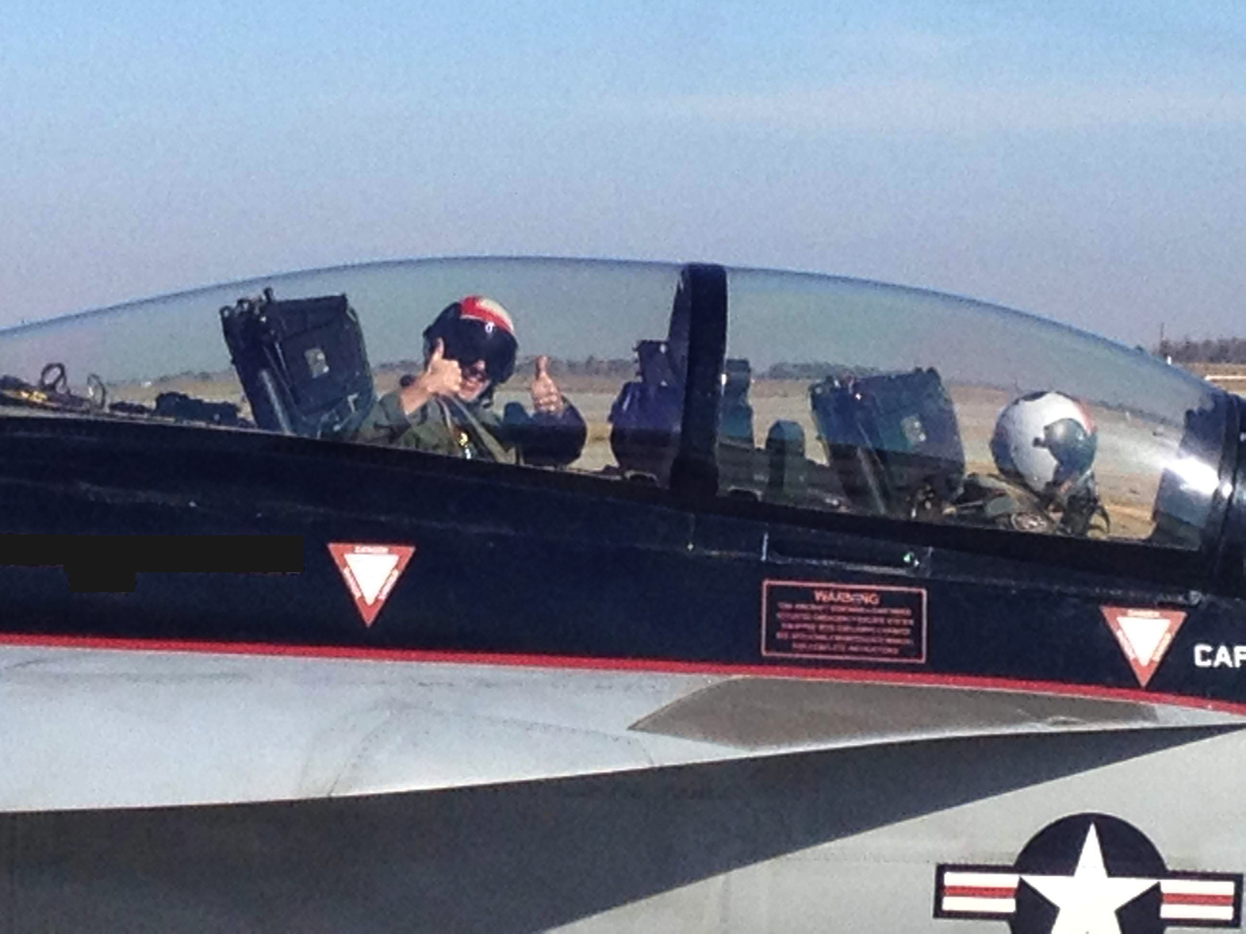 Pilot wearing a helmet and sunglasses giving a thumbs-up inside the cockpit of a fighter jet on an airfield.