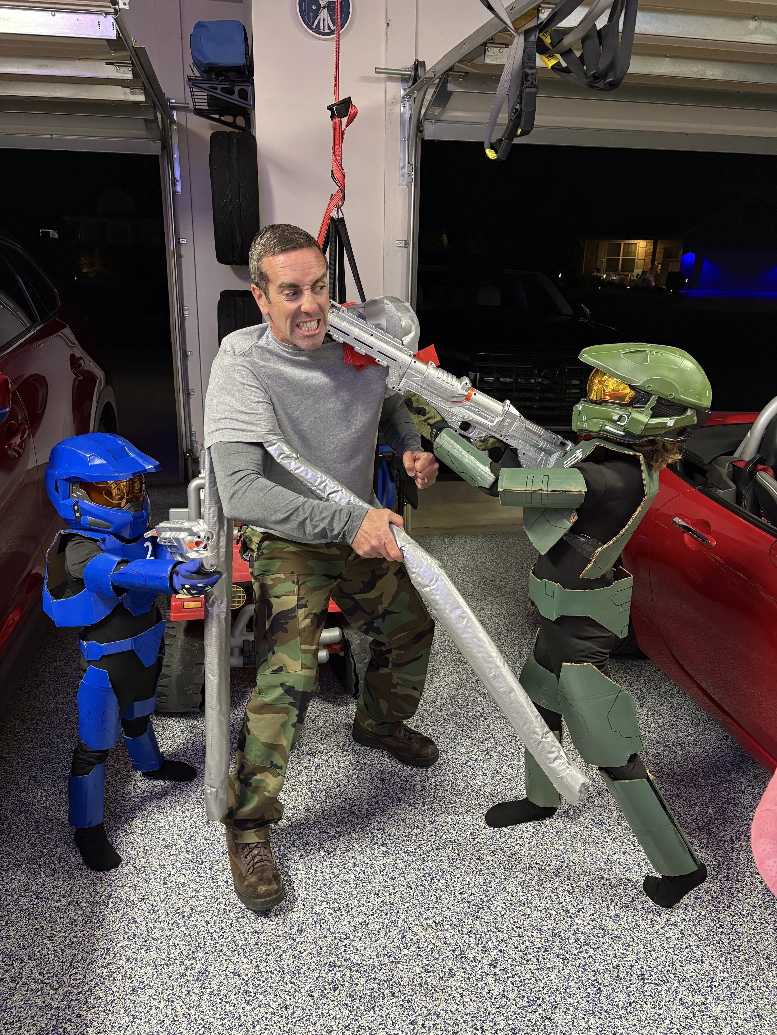 A man in camouflage pants fighting with life-sized cardboard cutouts of a blue armored soldier and a green armored soldier holding toy guns in a garage.