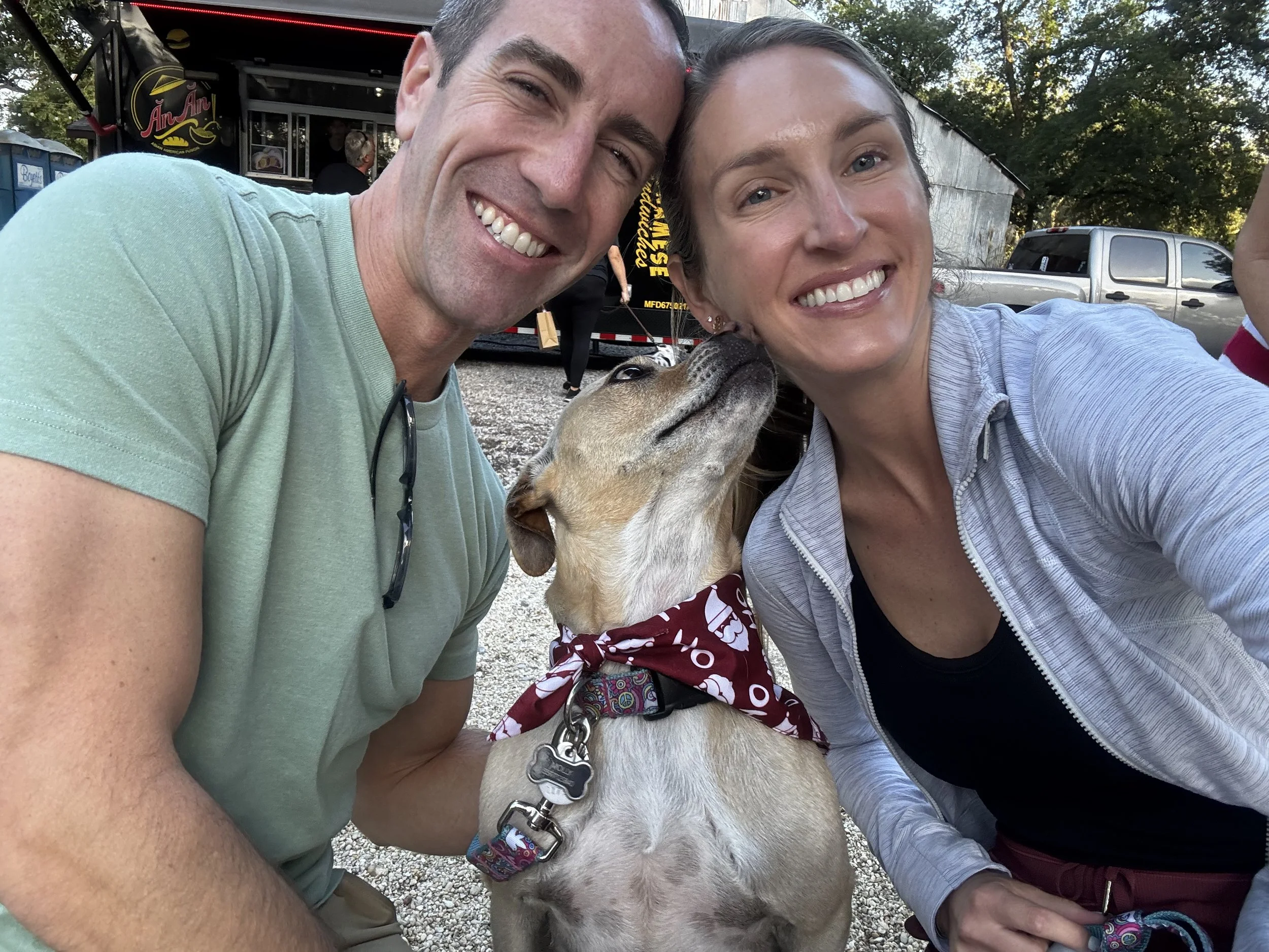 A smiling man and woman taking a selfie with their dog outdoors. The dog, wearing a maroon bandana, is licking the woman's face. There are trees and vehicles in the background.