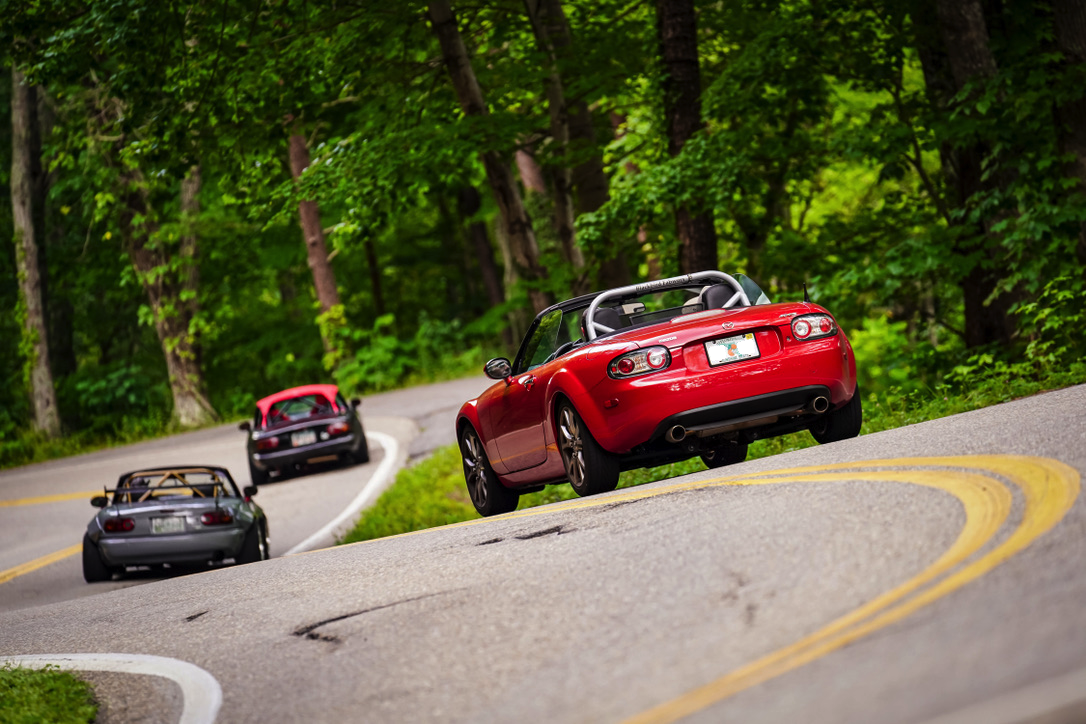 Four sports cars driving on a winding forest road with green trees in the background.