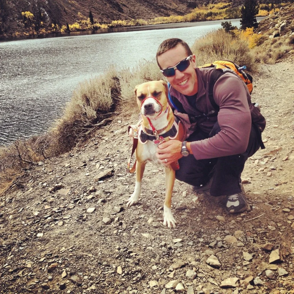 A man in sunglasses kneeling on a rocky trail by a river, smiling, with his dog. The landscape features a river, trees with fall foliage, and hills in the background.