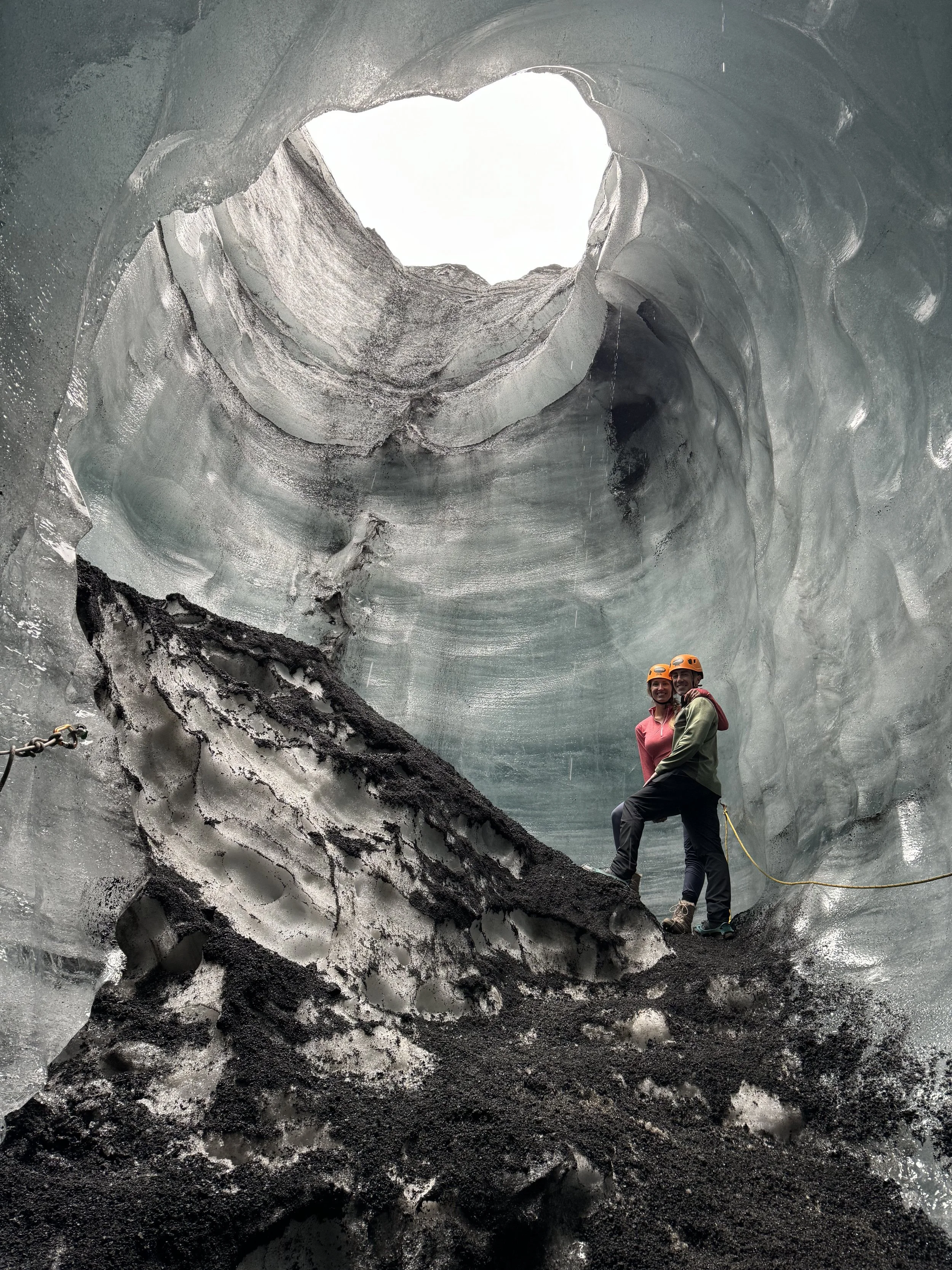 Two people wearing helmets standing inside a large ice cave with smooth, layered ice walls and a hole at the top letting in light.