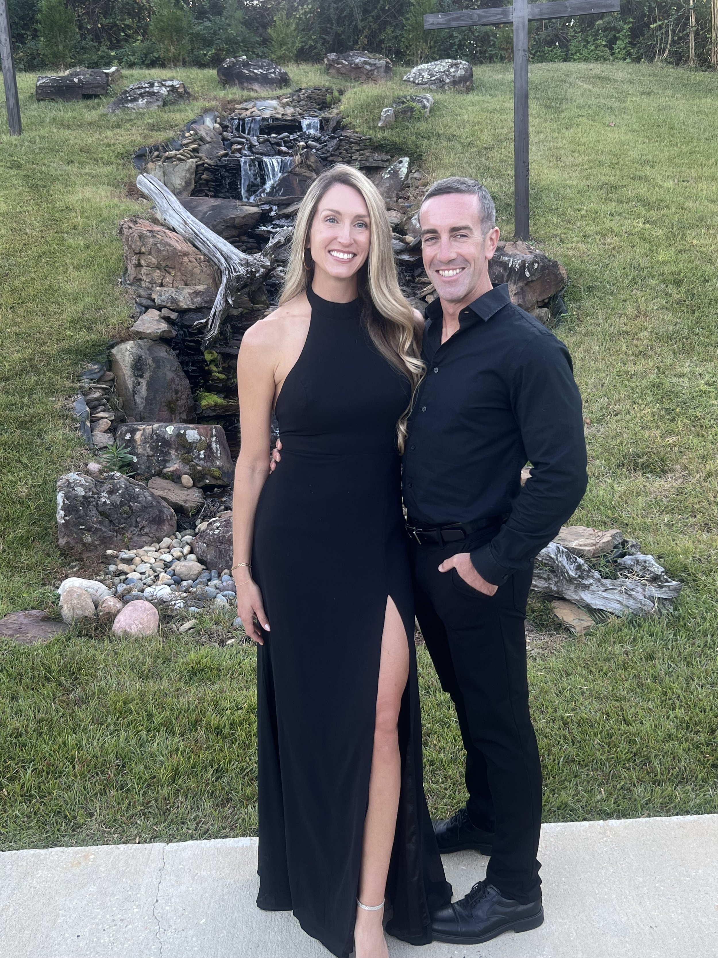 A smiling woman in a black dress with a high slit stands beside a smiling man in black dress clothes. They are outdoors on grass, with a stone waterfall feature and greenery in the background.
