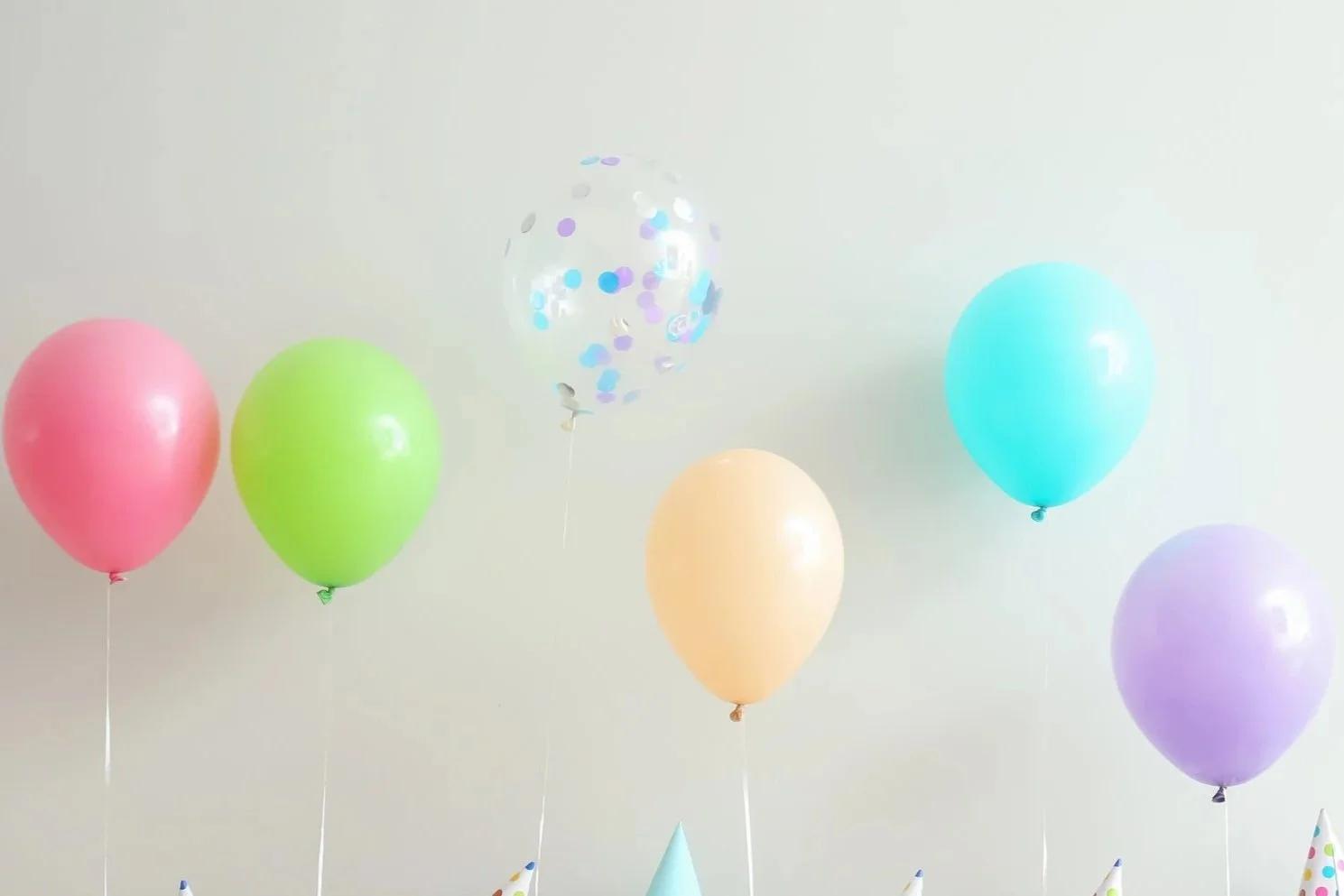 Colorful birthday balloons and party hats against a plain wall.