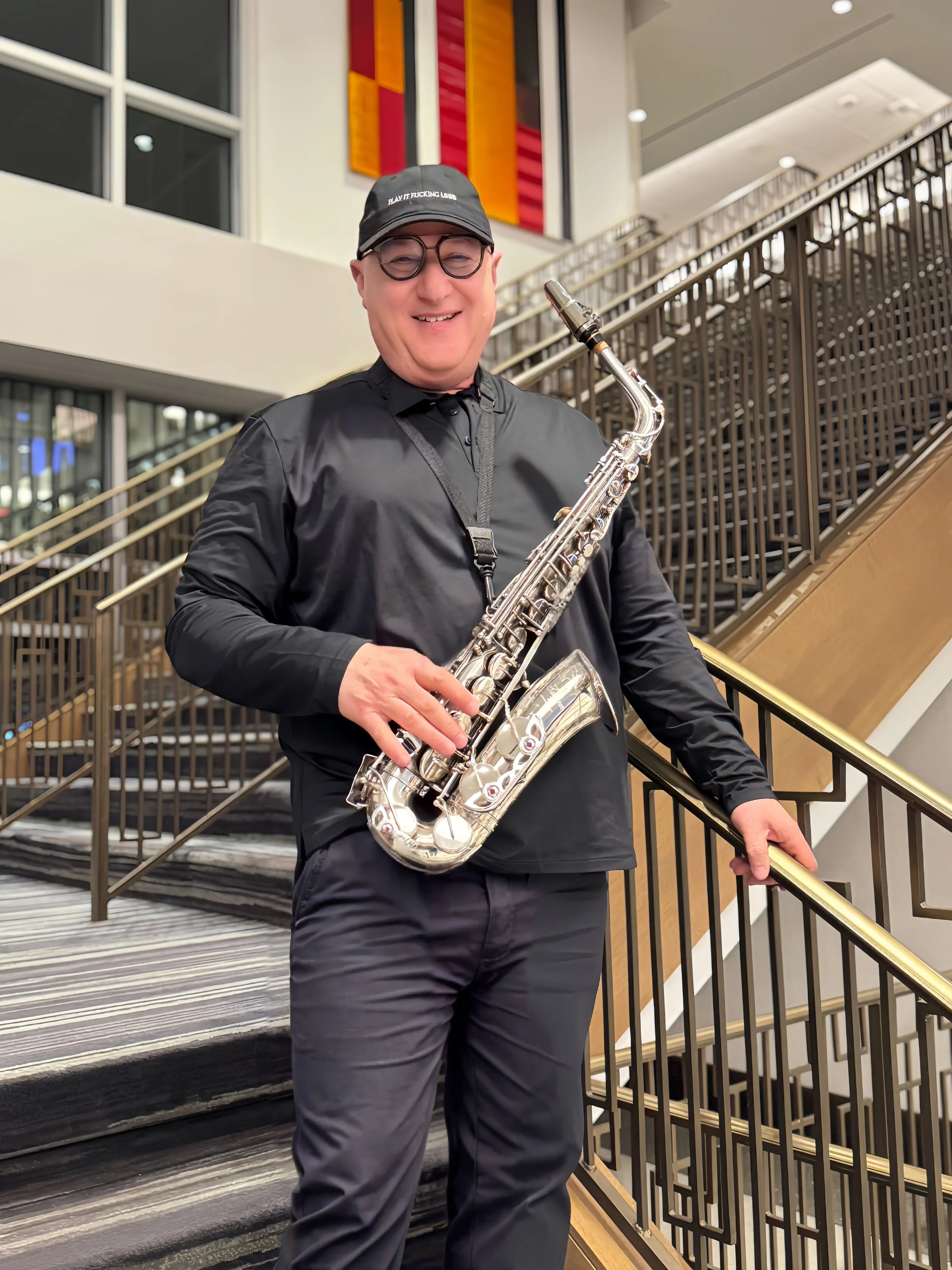 A smiling man wearing a black cap, glasses, and a black jacket, holding a silver saxophone, standing on a staircase in a modern indoor setting.