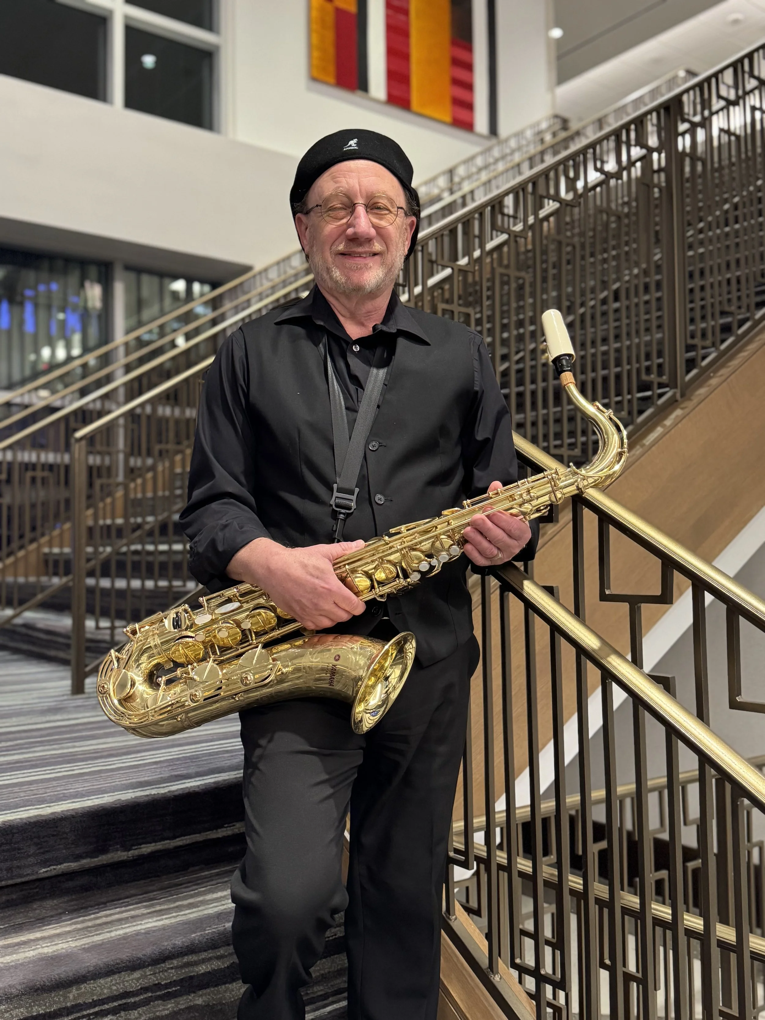 A smiling man dressed in black, wearing glasses and a black beret, holds a gold saxophone while standing on a staircase in an indoor setting.