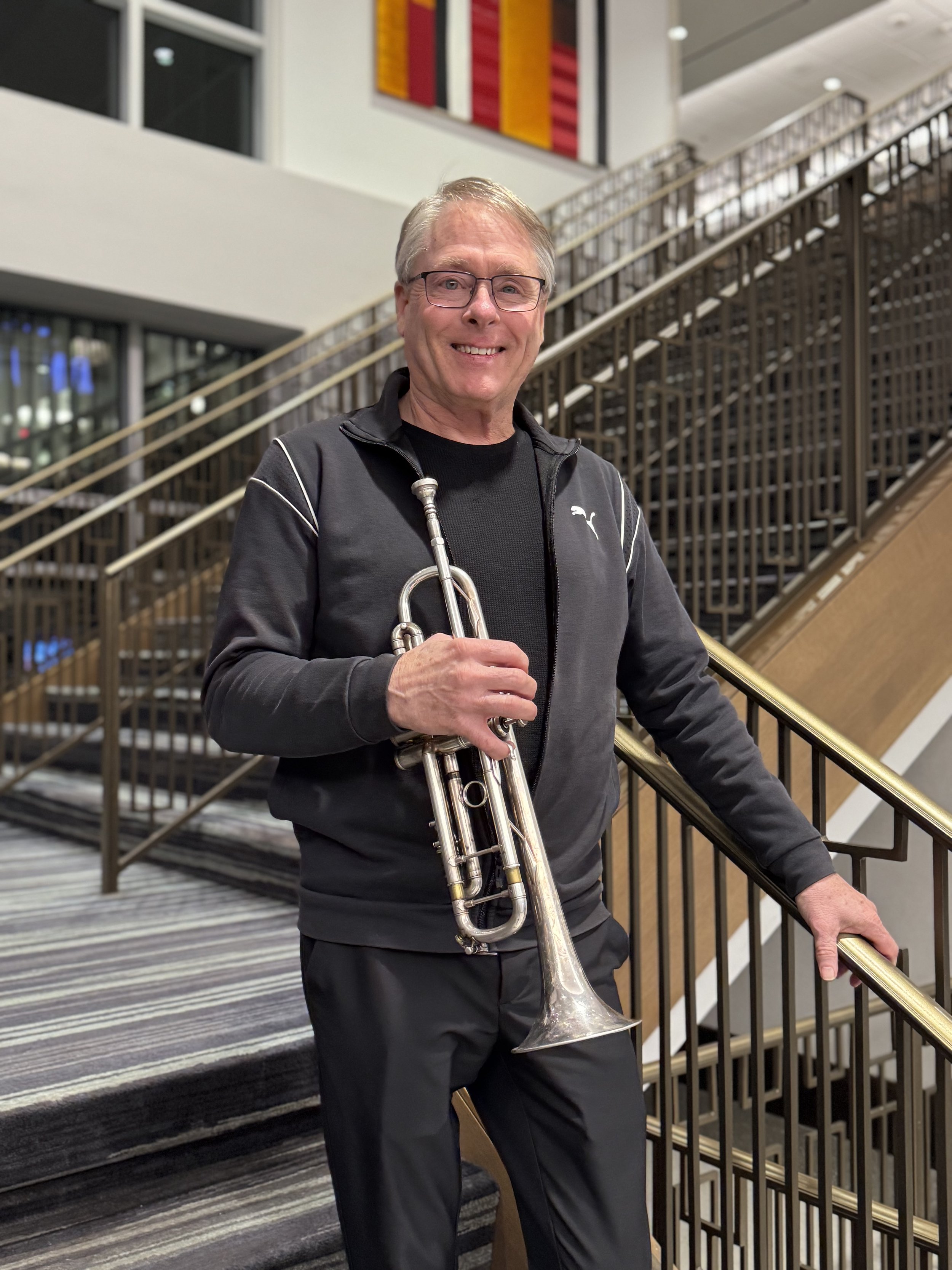 An older man with glasses, smiling, standing on a staircase inside a building, holding a trumpet in his right hand.