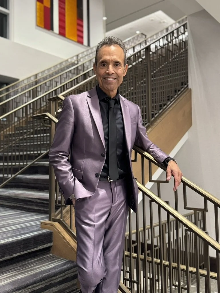 A man in a shiny lavender suit with a black shirt and tie, standing on a staircase in a modern interior building.