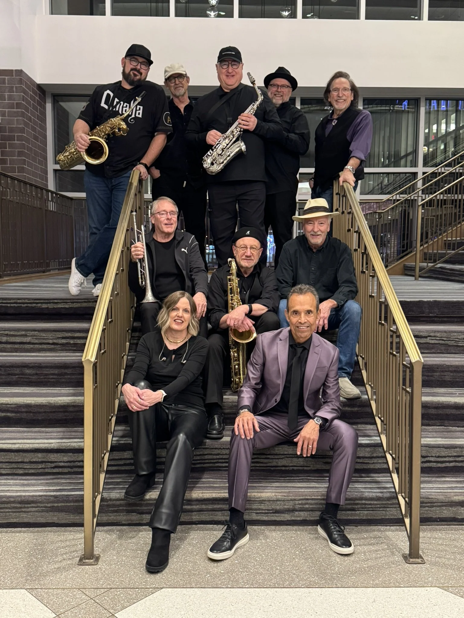 A group of ten funk musicians posing on a staircase inside a building, holding various musical instruments including saxophones, trumpet, and trumpet. They are smiling and dressed in casual and semi-formal attire.