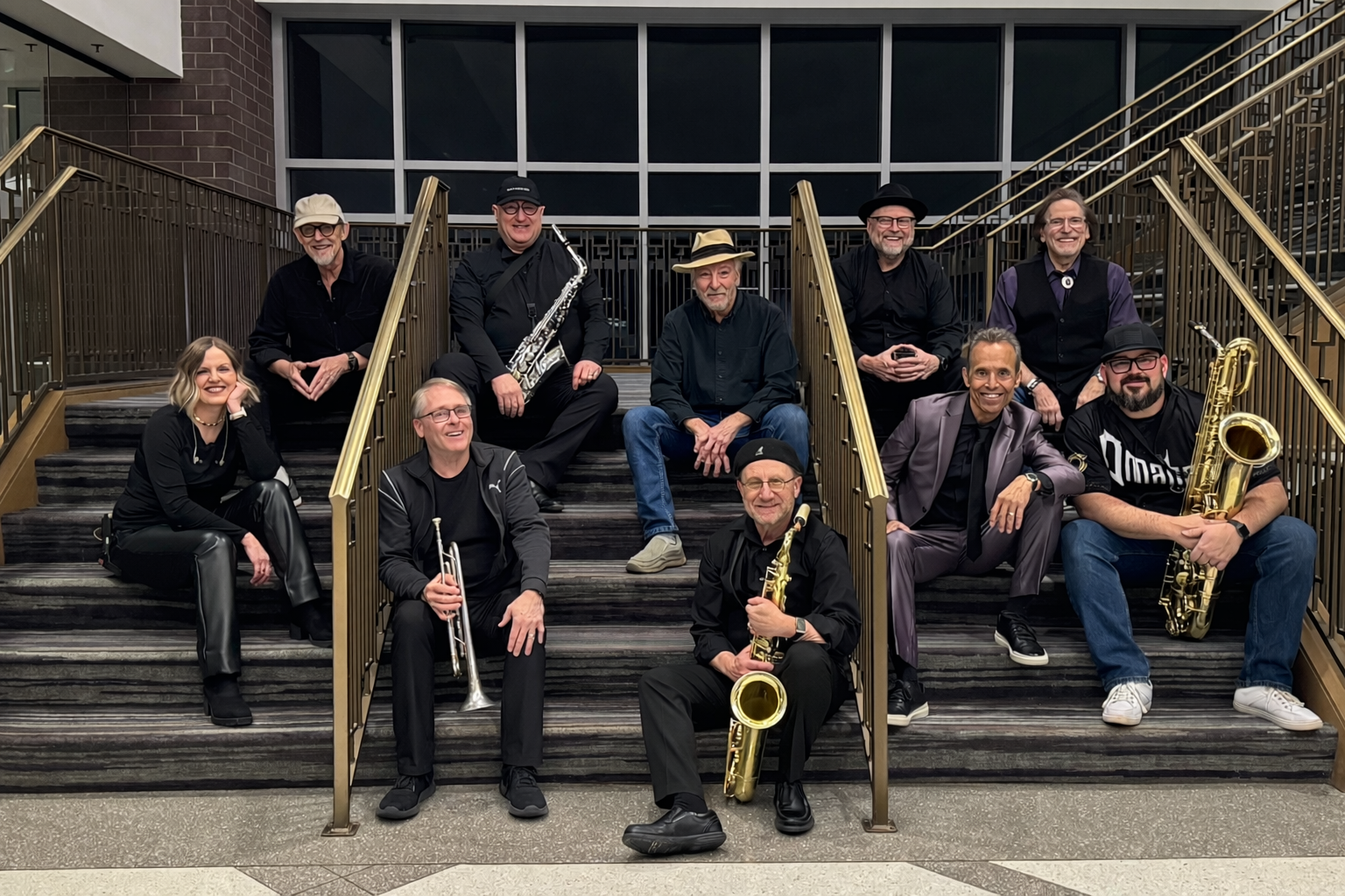 A group of ten people, some holding musical instruments like saxophones and trumpet, posed on a staircase in an indoor setting.