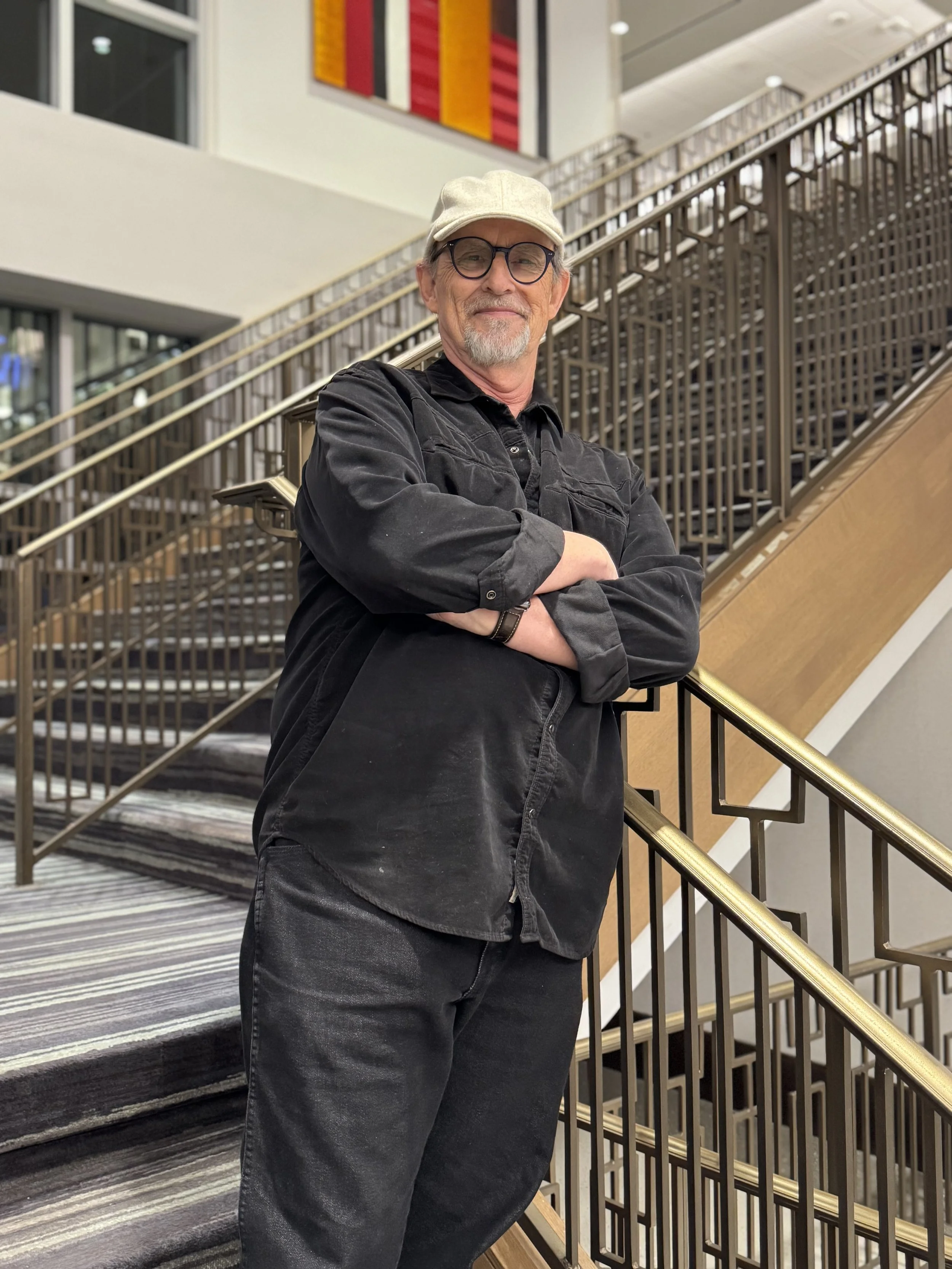 A man with a beard and glasses wearing a beige cap, black shirt, and dark pants, standing on a staircase with his arms crossed indoors.