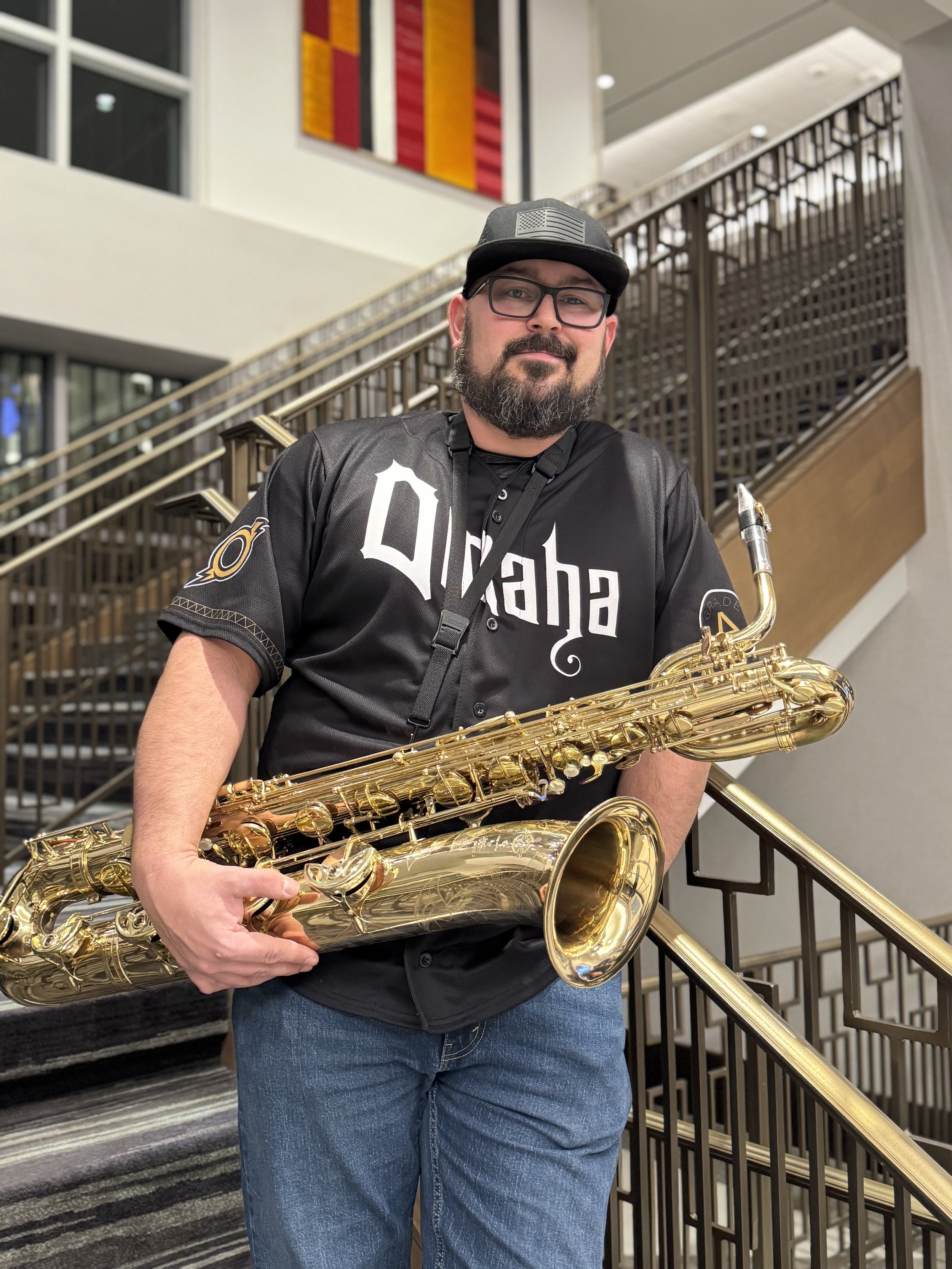 A man with a beard and glasses, wearing a black baseball cap and a black jersey with 'Duh' written on it, holding a gold saxophone, standing on a staircase in a modern indoor setting.