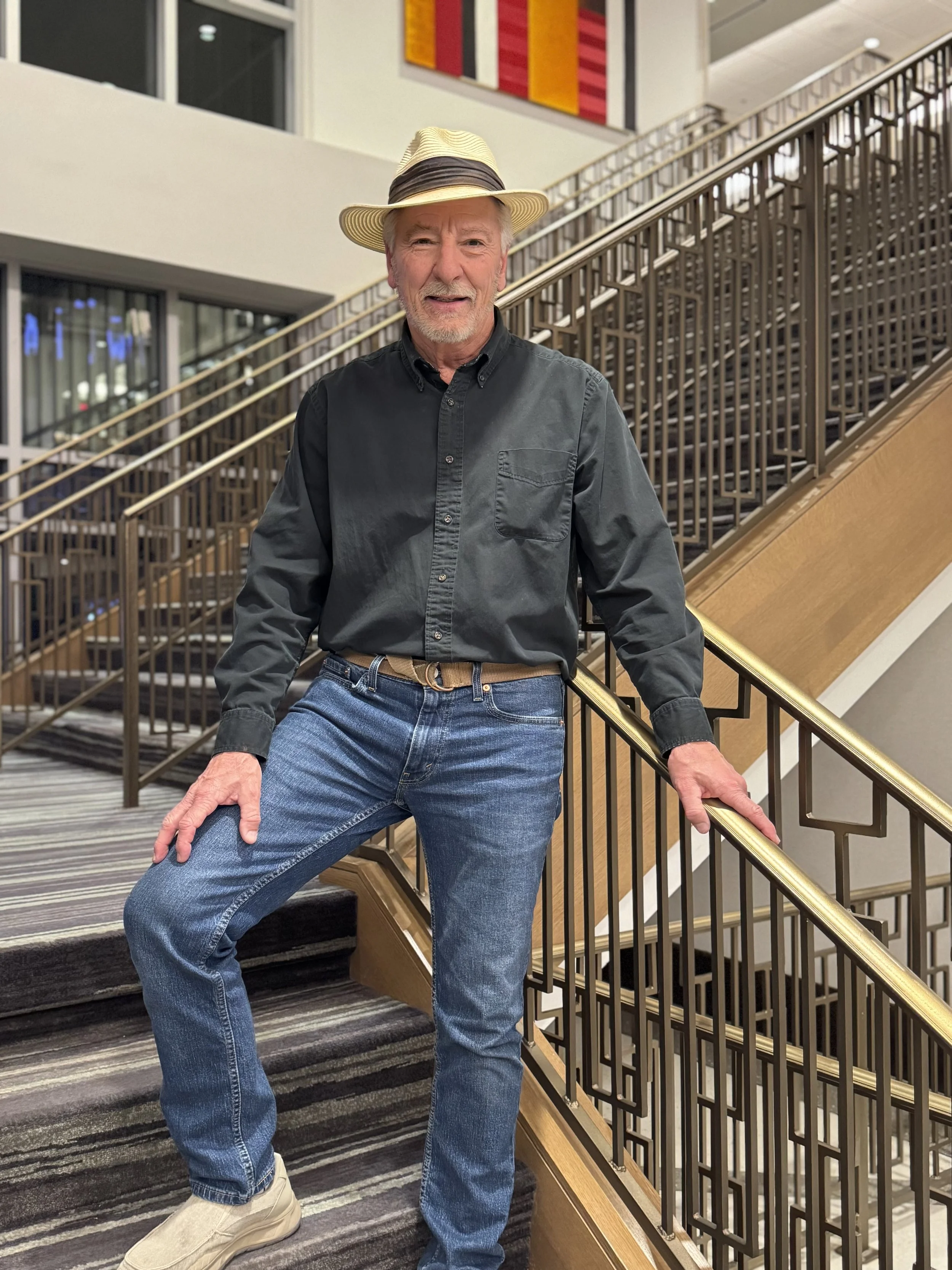 An older man wearing a striped straw hat, black button-up shirt, blue jeans, and light-colored shoes, standing on a carpeted staircase inside a modern building with a railing and colorful artwork on the wall behind him.
