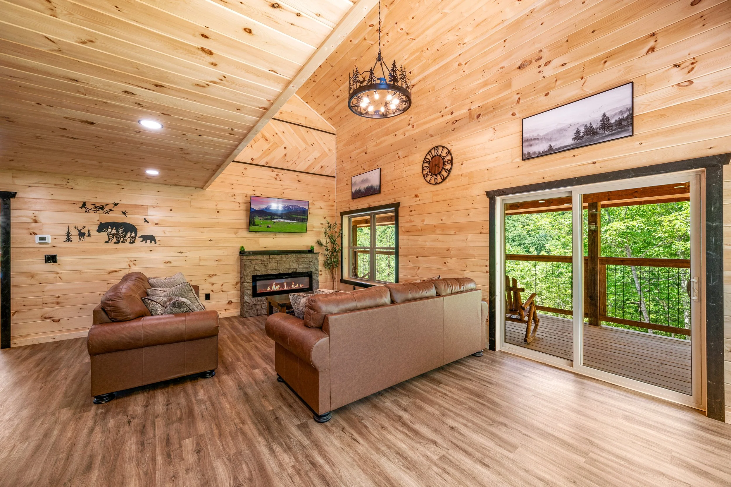 Living room with wood-paneled walls and ceiling, two brown sofas, a fireplace, mounted TV, wall art, large windows, and a sliding glass door leading to a balcony with a view of green trees.