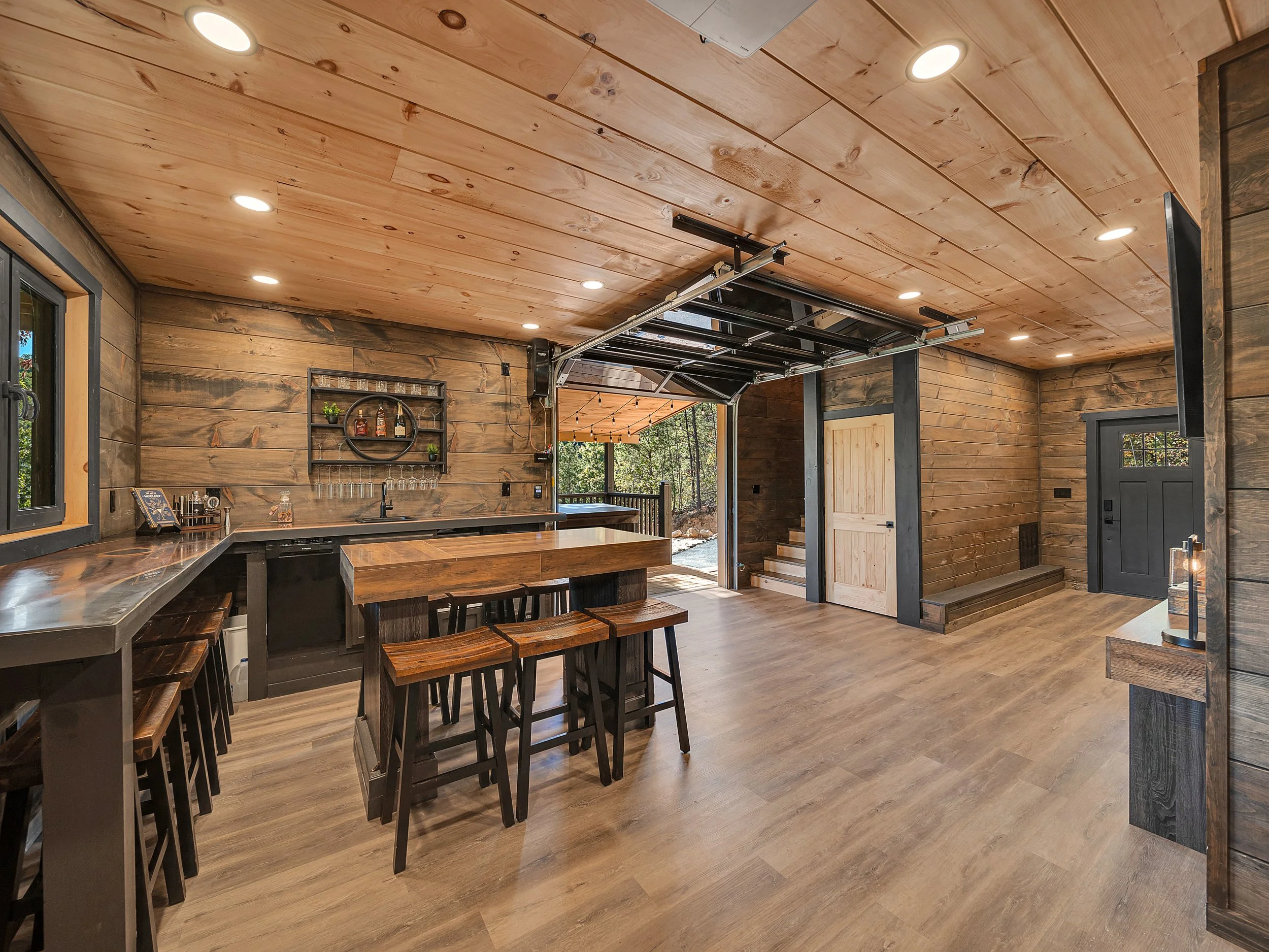 Interior of a rustic bar or kitchen area with wood-paneled walls and ceiling, a small bar counter, bar stools, and a garage-style door open to an outdoor space with trees.