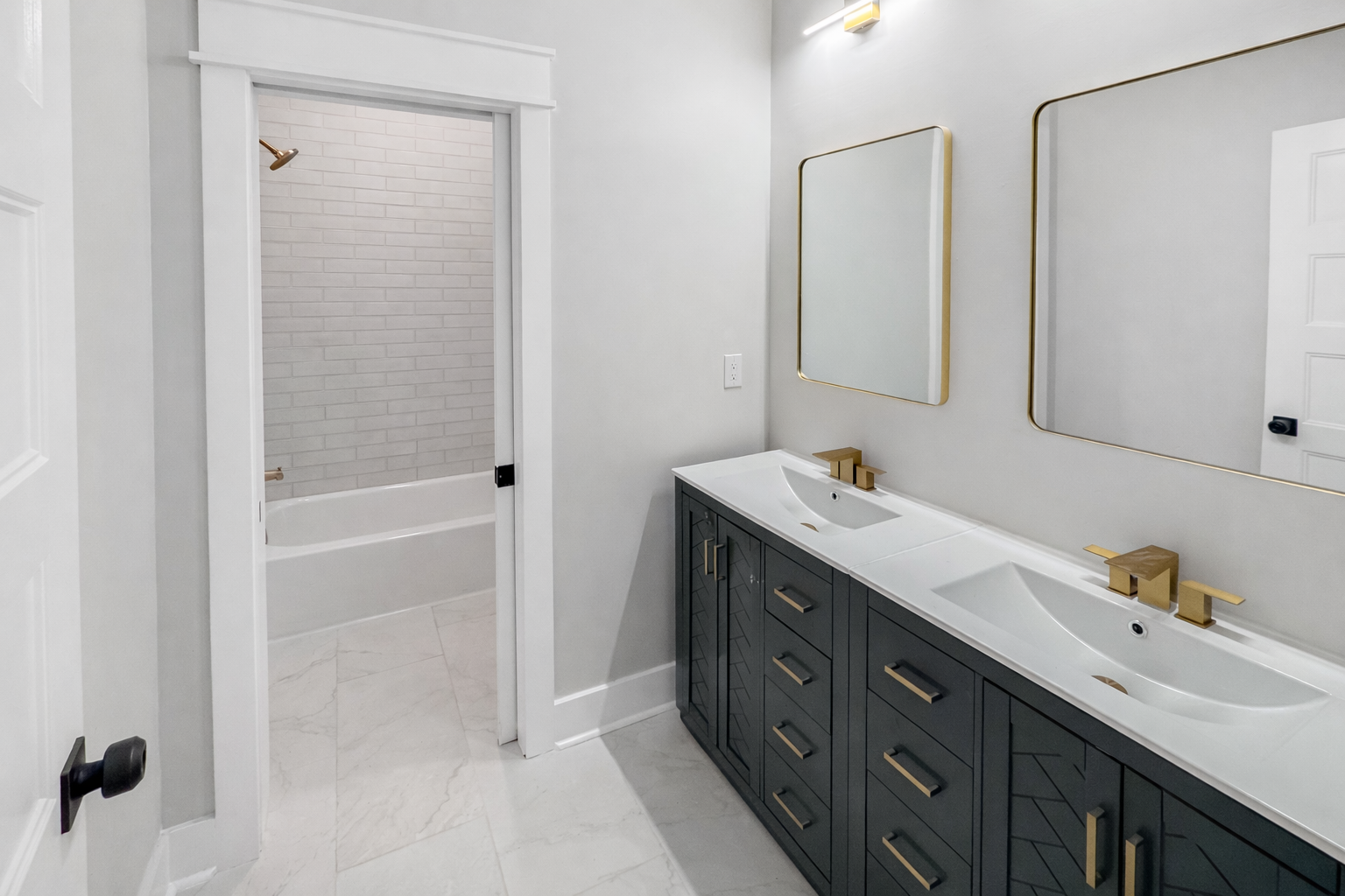 Modern bathroom with double vanity sink, gold fixtures, and two large mirrors. In the background, there is a bathtub with a shower area that has white brick tiles. The bathroom features white walls, light-colored marble flooring, and a door with a bl
