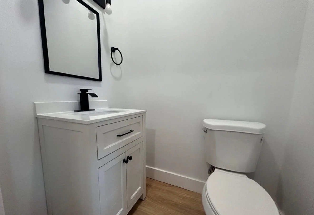 A small, modern bathroom with white walls and a light wood floor. It features a white vanity with a black faucet, two black knobs, and a small rectangular mirror above. To the right, there is a white toilet. There is a black towel ring mounted on the