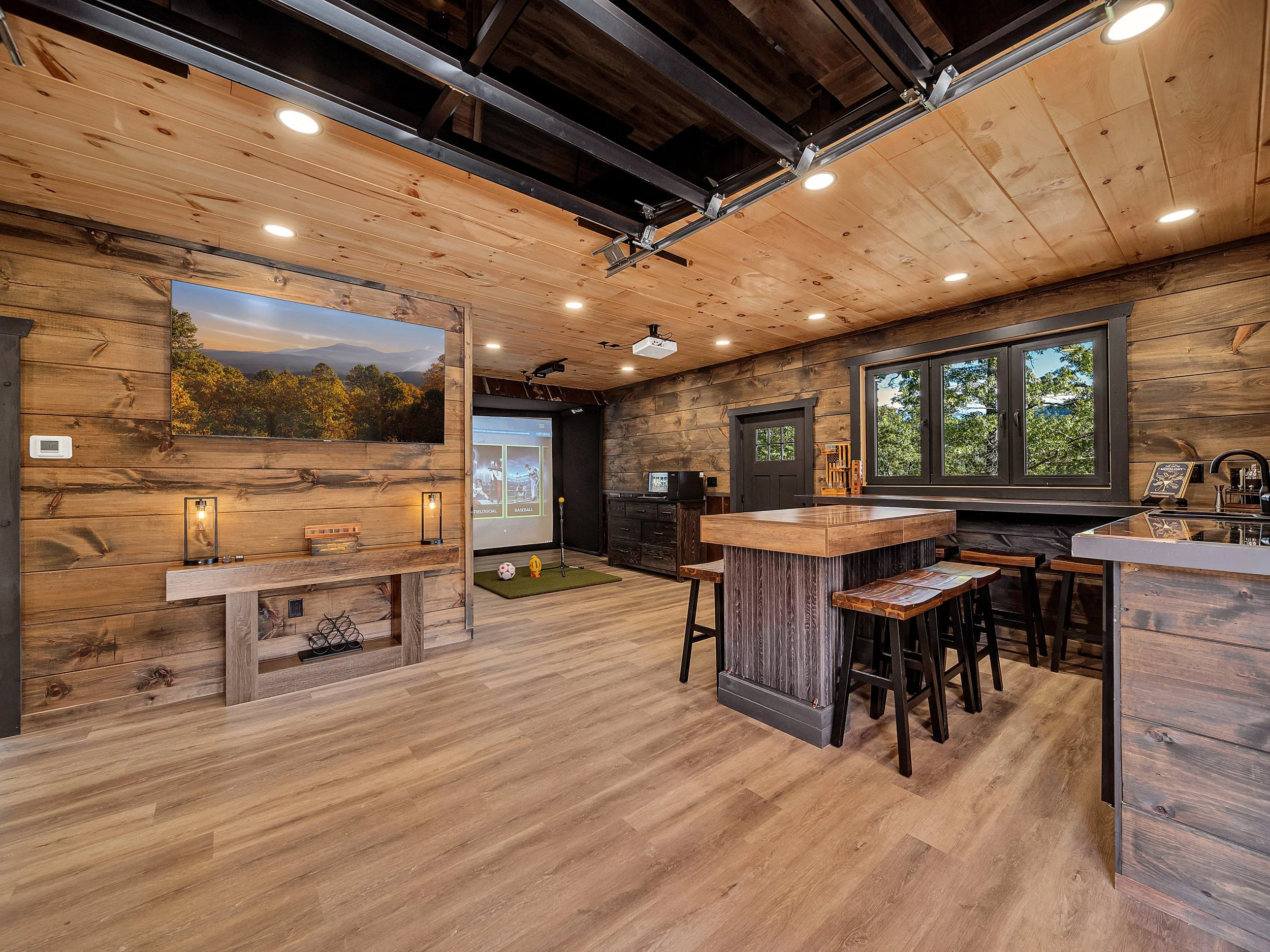 Interior of a rustic living space with wooden walls, ceiling, and flooring. Features a mounted large landscape photo, a TV, a projector, a pool table, and a kitchen area with a window and bar stools.