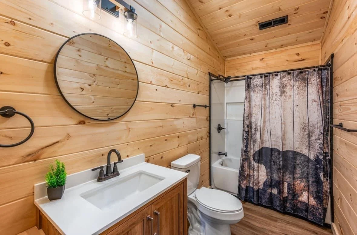 Bathroom with wood-paneled walls, a round mirror, a small plant on the sink, a toilet, and a shower with a curtain featuring a bear design.
