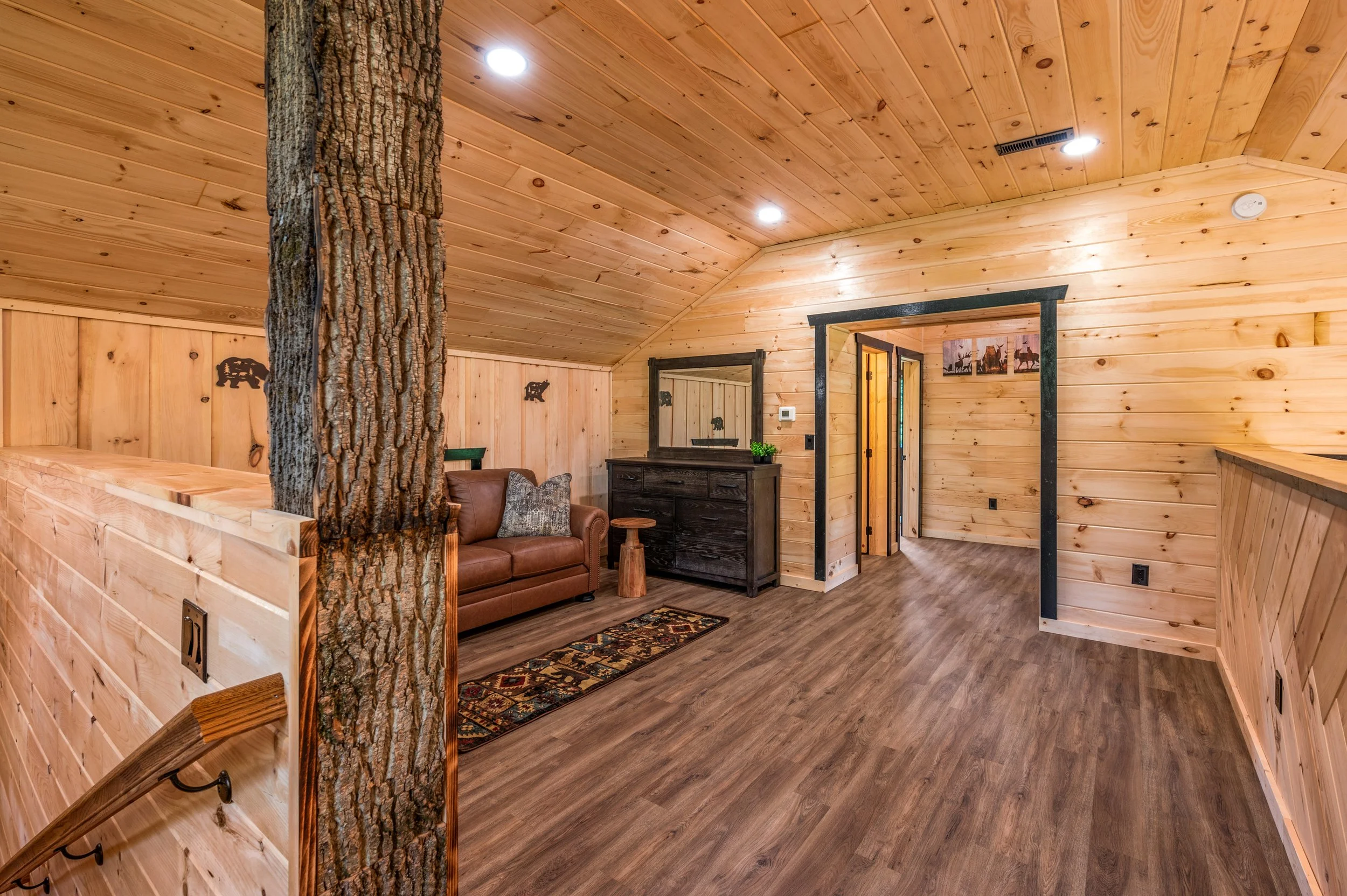 Interior of a log cabin-style room with wood-paneled walls and ceiling, a faux tree trunk, a brown leather sofa with patterned pillows, a black wooden dresser with a mirror, a small wooden stool, framed wildlife pictures, and a wooden floor.