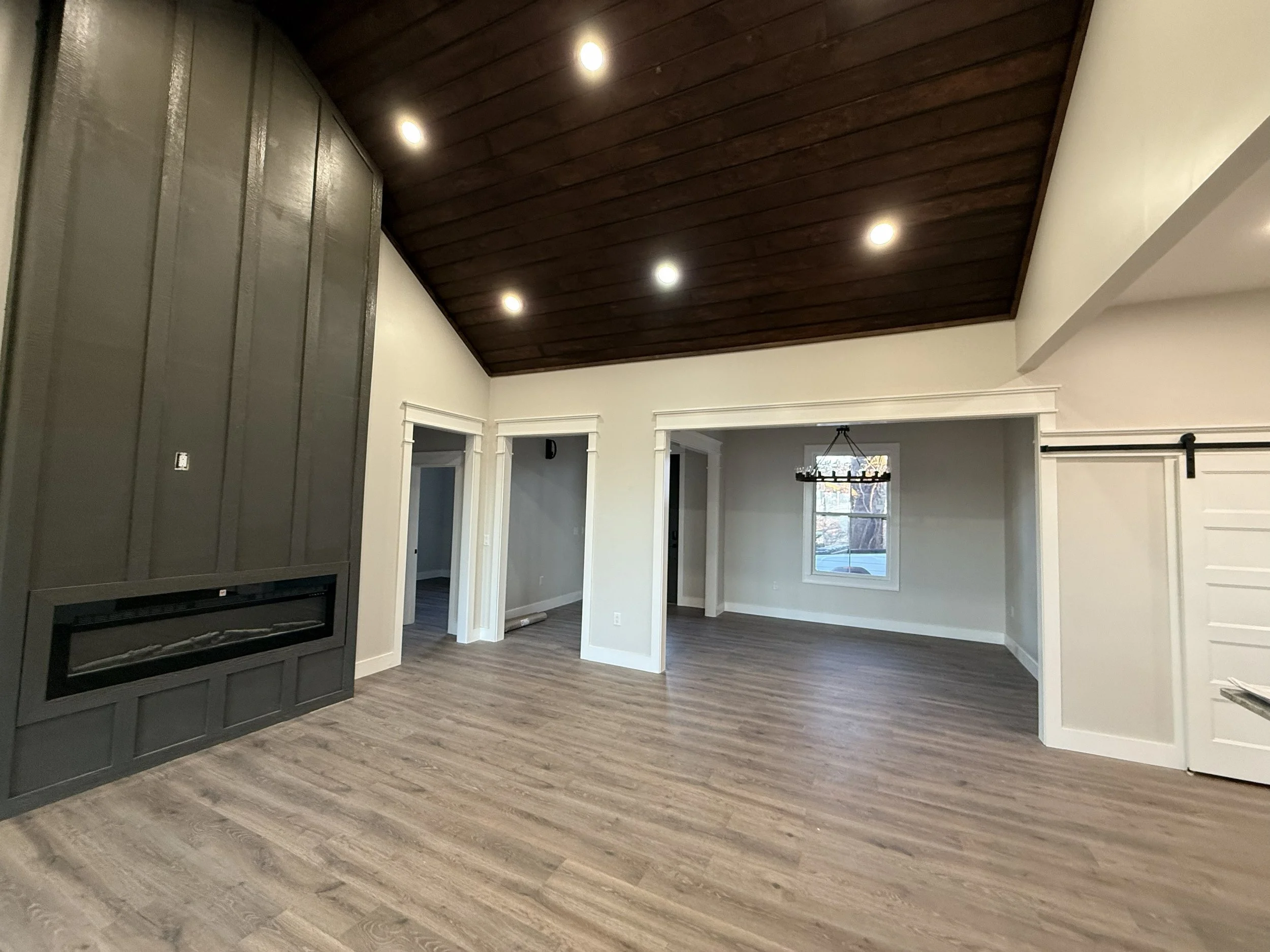 Interior view of a living room with wood floors, white walls, and a dark wood vaulted ceiling. There is a gray accent wall with a built-in fireplace. The room has multiple doorways and a window with a chandelier hanging in the adjacent room.