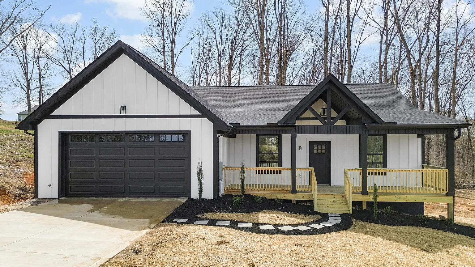 Newly constructed modern house featuring a black and white exterior, black garage door, small porch with wooden railing, and a driveway, set in a wooded area.