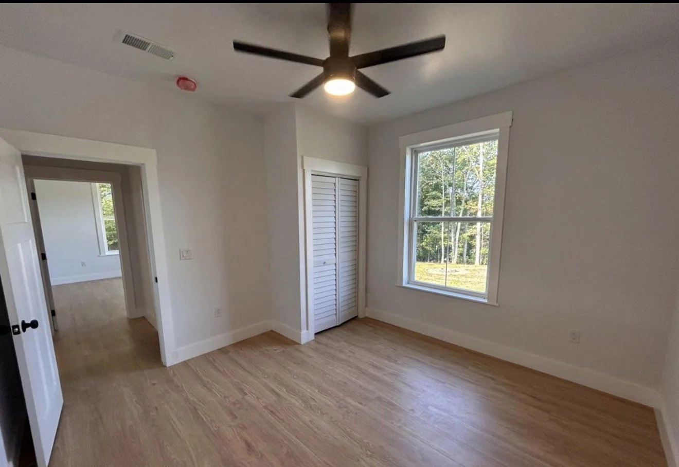 A bright, empty bedroom with a large window overlooking greenery, a closet with louvered doors, a ceiling fan with lights, white walls, and hardwood floors.