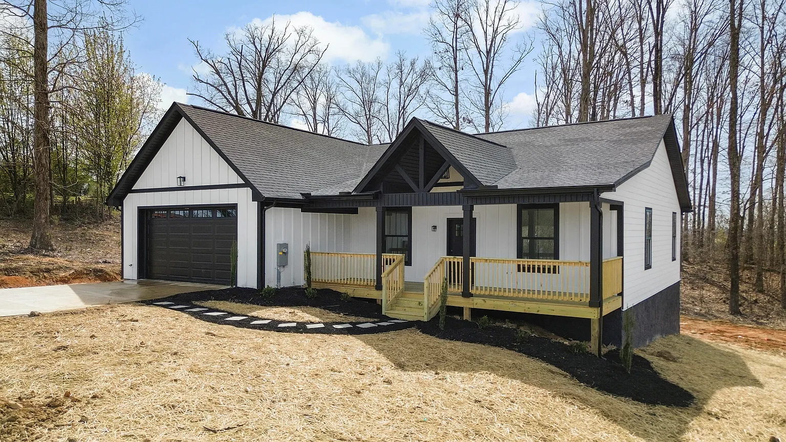 Newly constructed modern white house with black accents, attached garage, front porch with wooden railing, surrounded by trees and a cleared yard.
