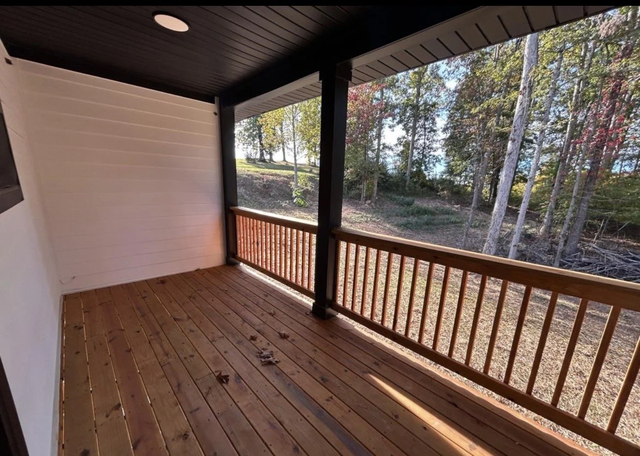 Empty wooden porch with railing overlooking a wooded yard with trees and sunlight.