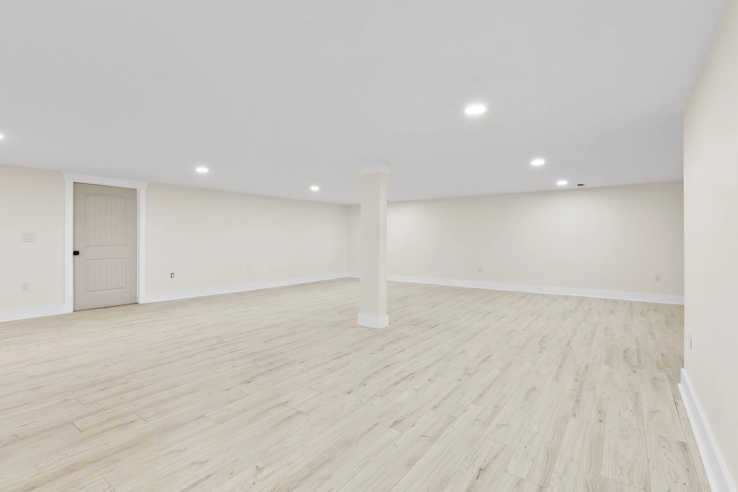 Empty room with white walls, white ceiling with recessed lighting, light wood flooring, and a closed beige door.