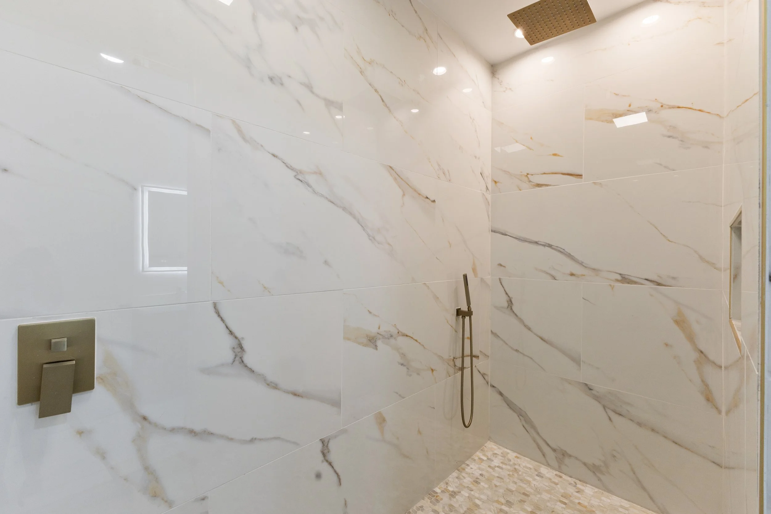 Walk-in shower with large white marble tiles, a rain showerhead, and a handheld shower fixture. Recessed ceiling lights and a small built-in niche for toiletries.