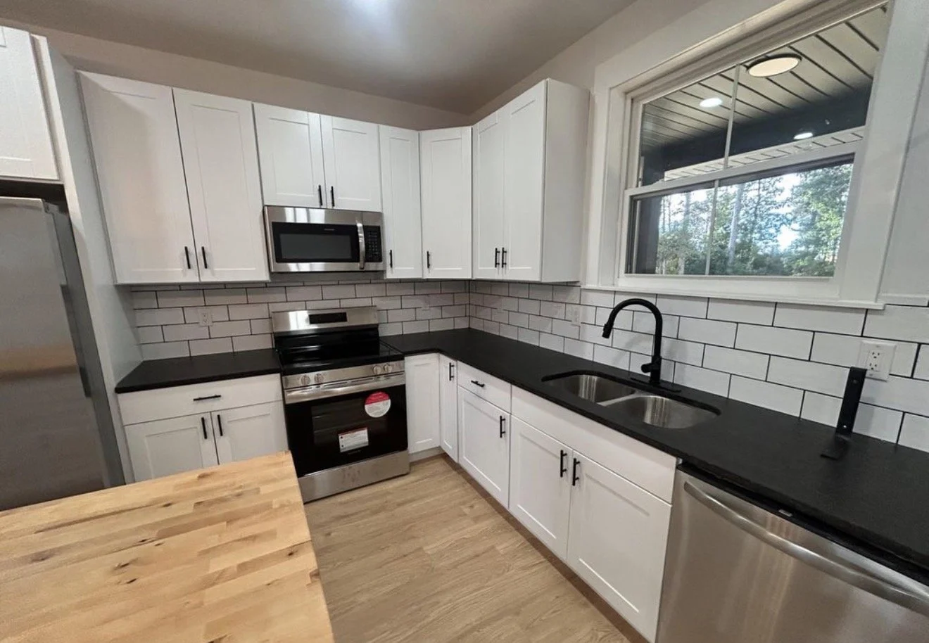 Modern kitchen with white cabinets, black countertops, stainless steel appliances, a window above the sink, and a wooden dining table.