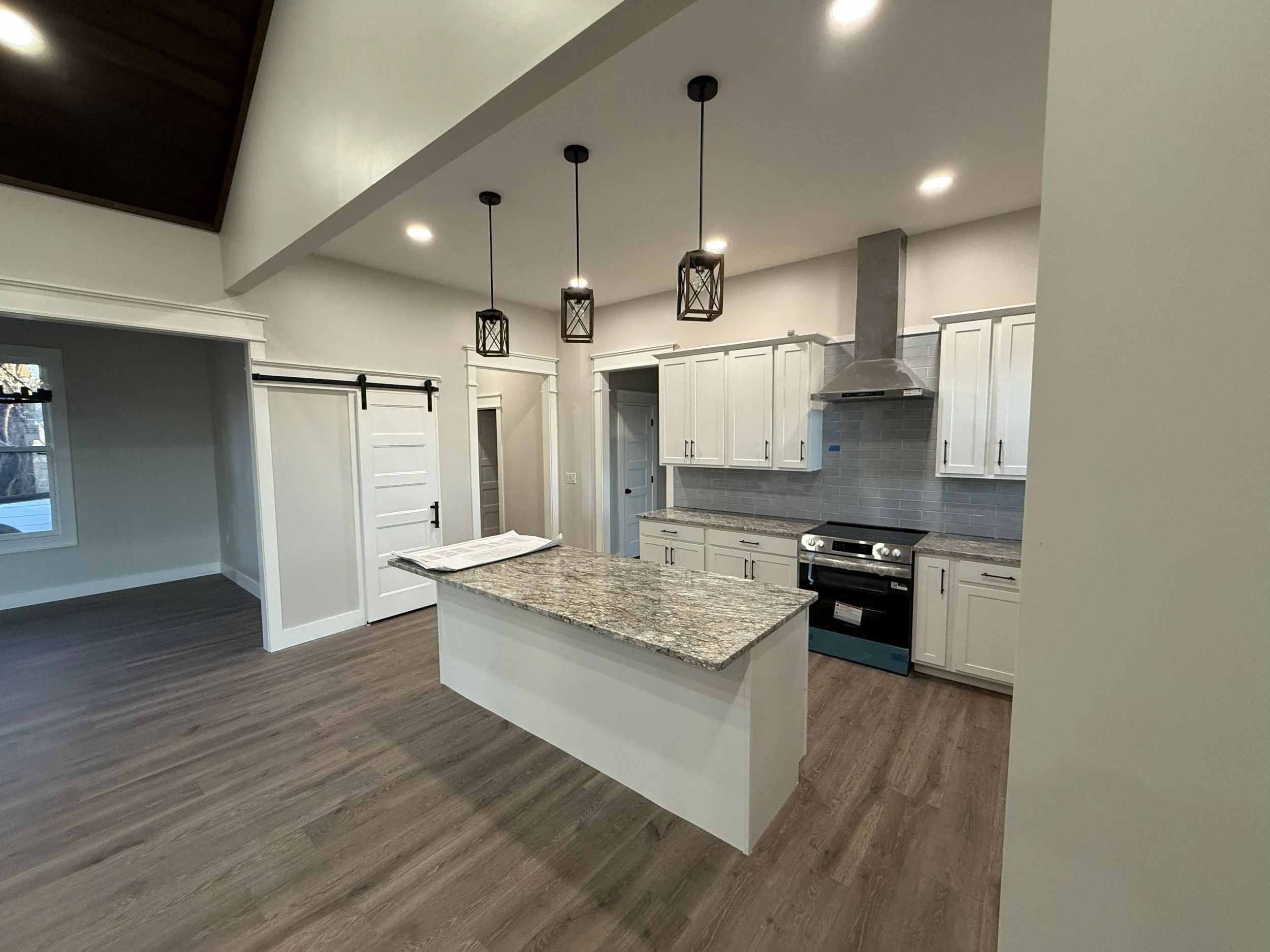 Modern kitchen with white cabinets, granite countertop island, stainless steel stove, gray tile backsplash, hanging pendant lights, and wood flooring.