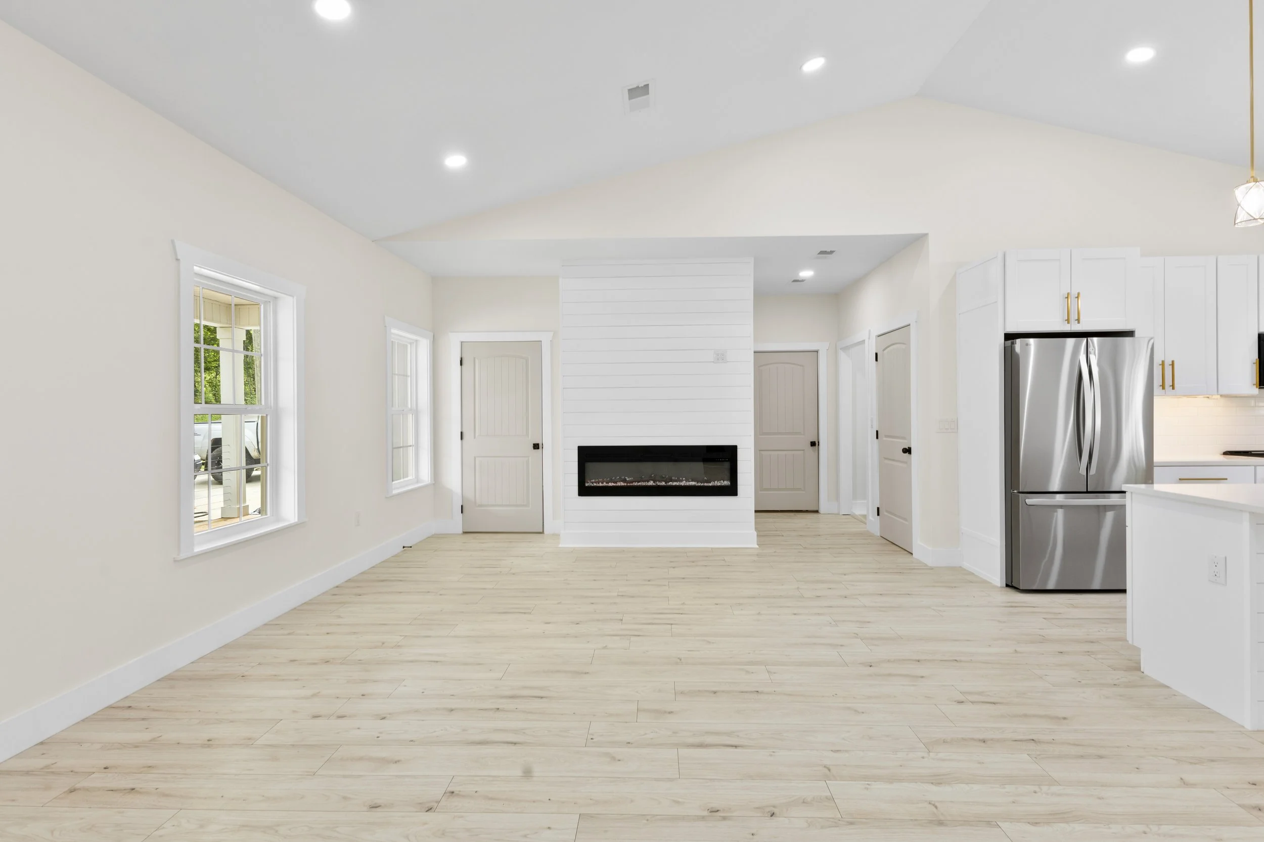 Empty living room with white walls, light wooden flooring, and a modern fireplace on a white wall with horizontal paneling. There are three windows on the left, with a view of a porch and green trees outside. The ceiling is vaulted with recessed ligh