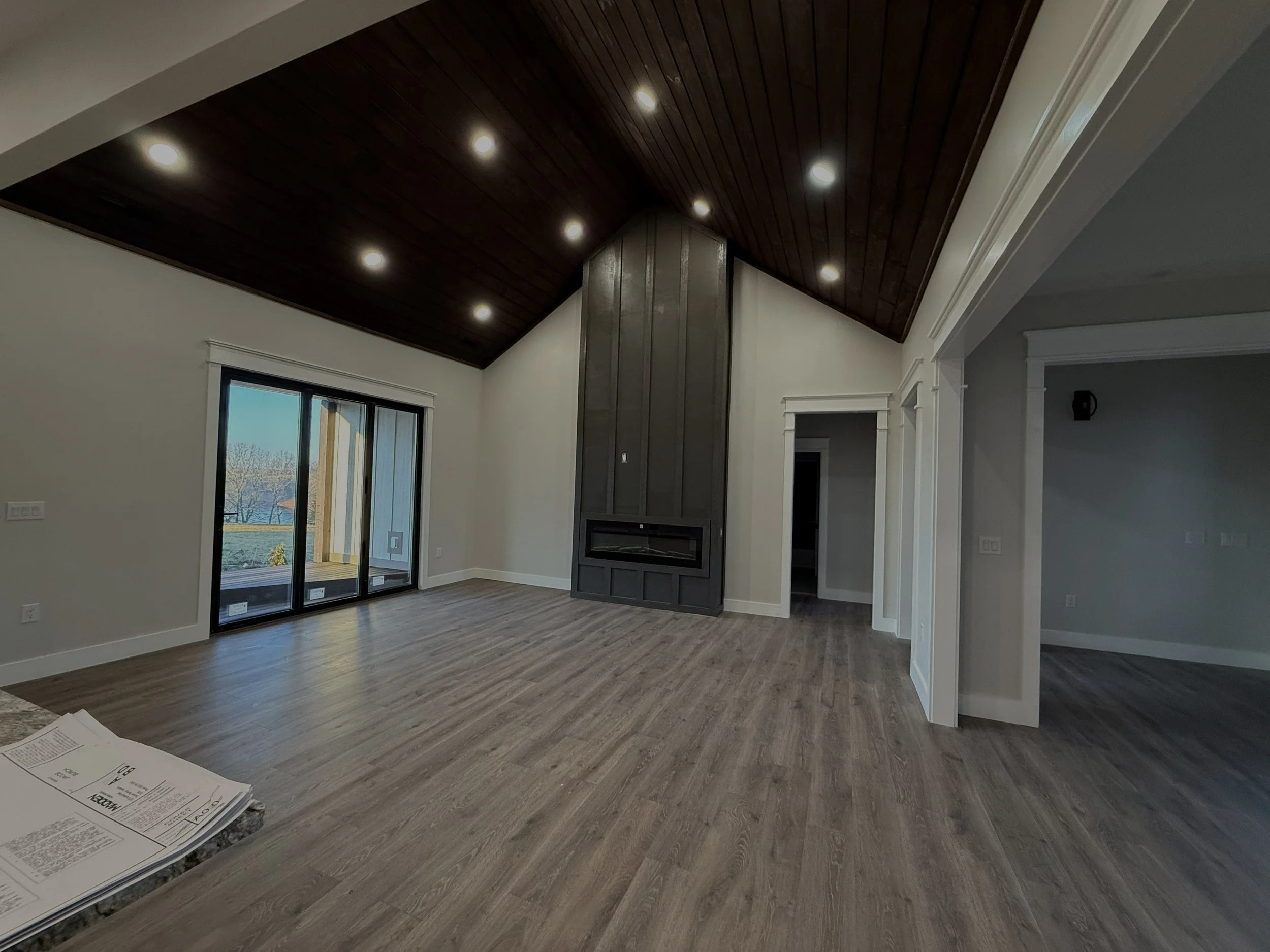 Empty living room with wooden floor, vaulted dark wood ceiling with recessed lights, white walls, large sliding glass door leading to outdoor area, and a built-in dark wood fireplace.