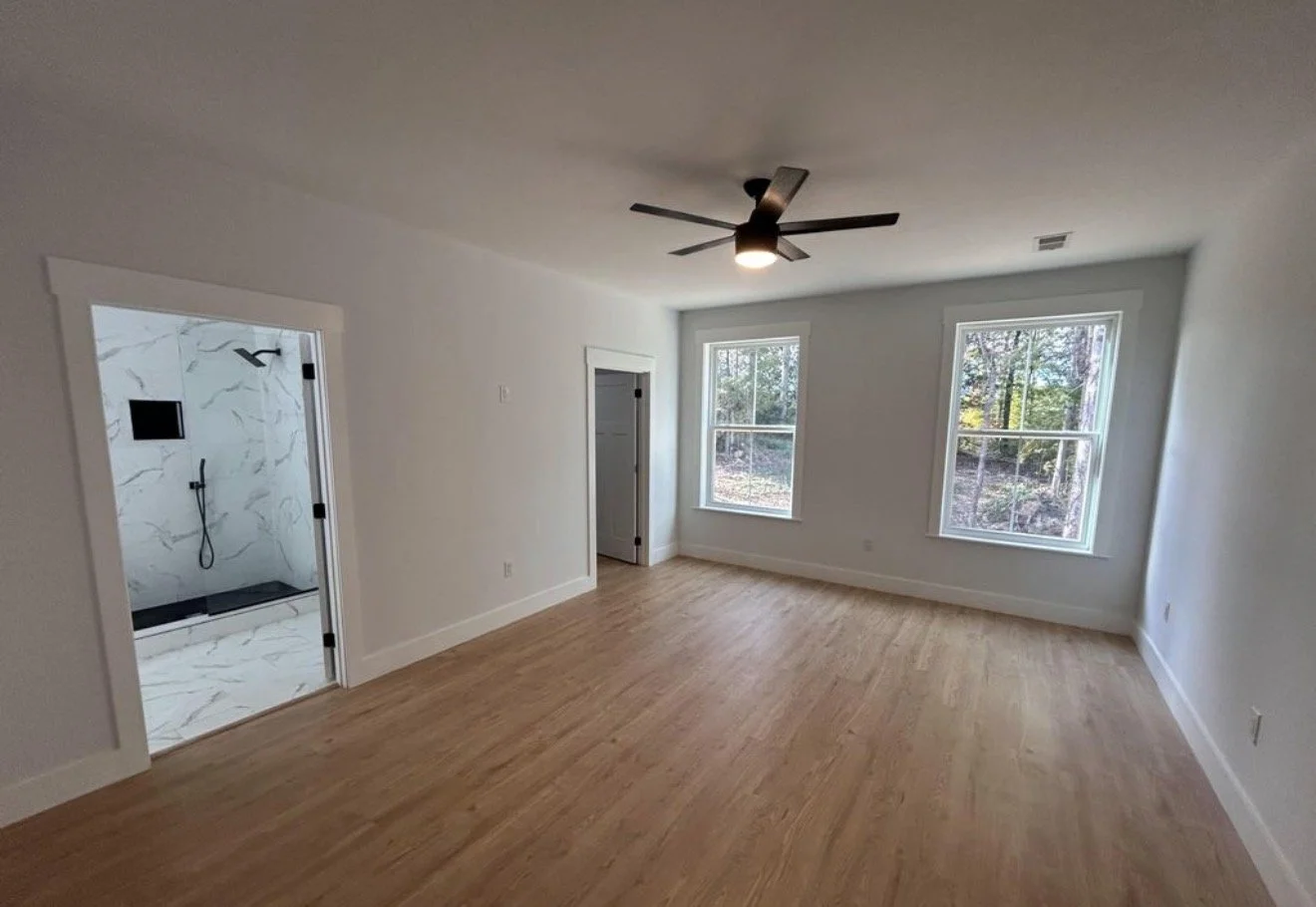 Empty bedroom with light wood flooring, white walls, two large windows showing trees outside, ceiling fan, and an open door leading to a bathroom with marble tile shower.