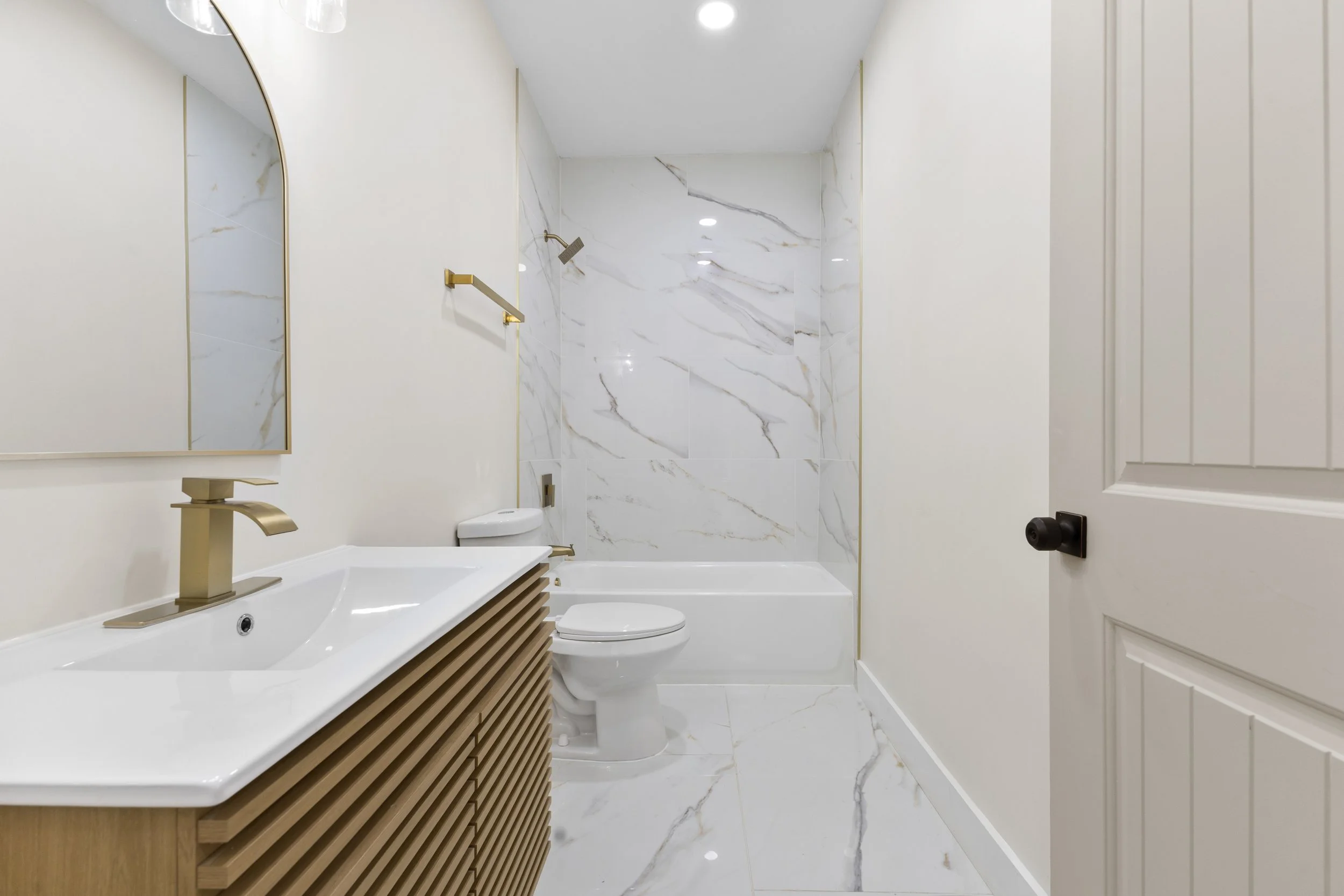 Modern bathroom with white marble walls and floors, a wooden vanity with a white sink, a gold faucet, a mirror, a toilet, a bathtub with a shower, and a door, with ceiling lights