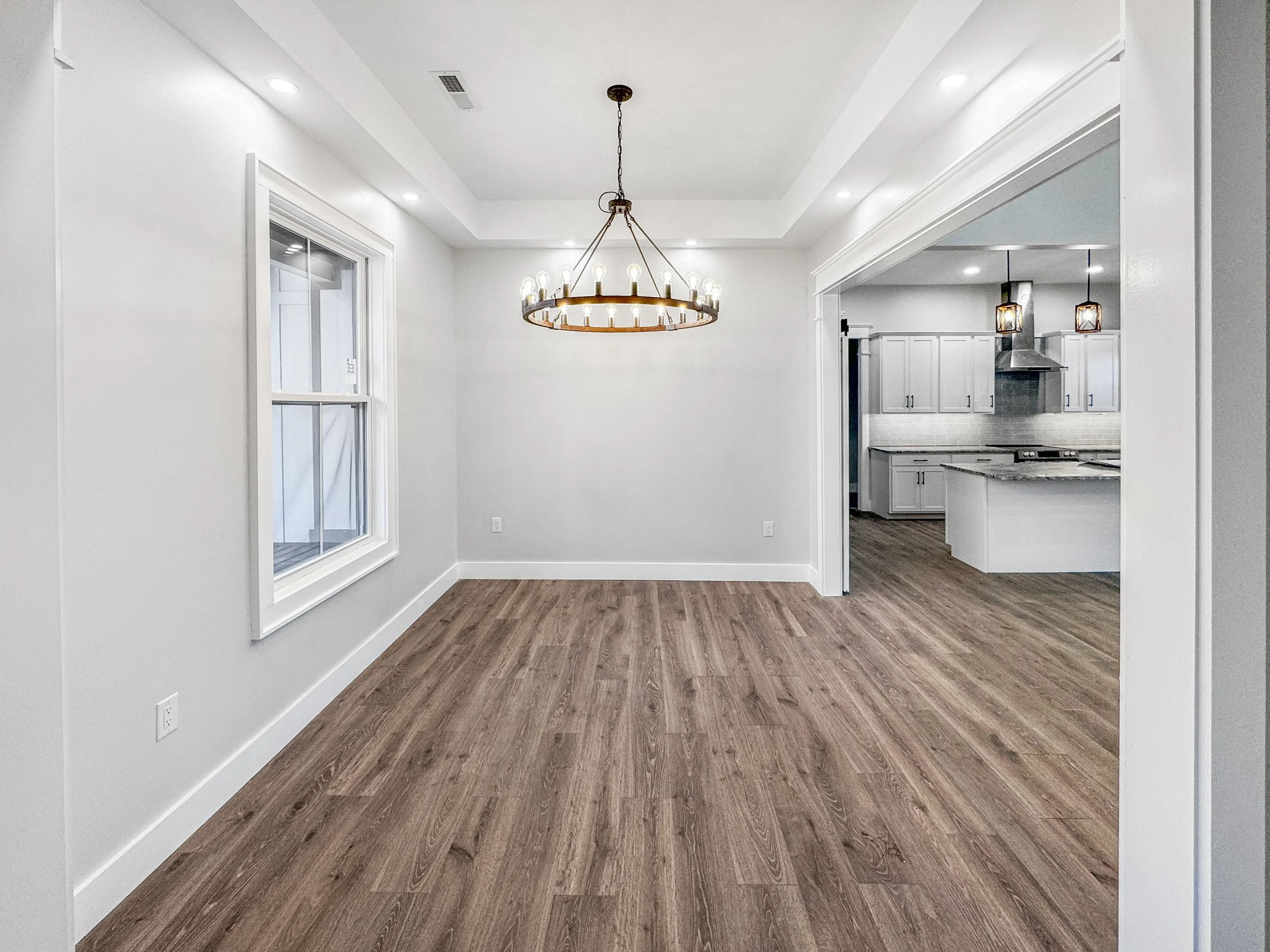 Empty dining room with hardwood floors, white walls, a chandelier, and a view into the kitchen with white cabinets and modern lighting.