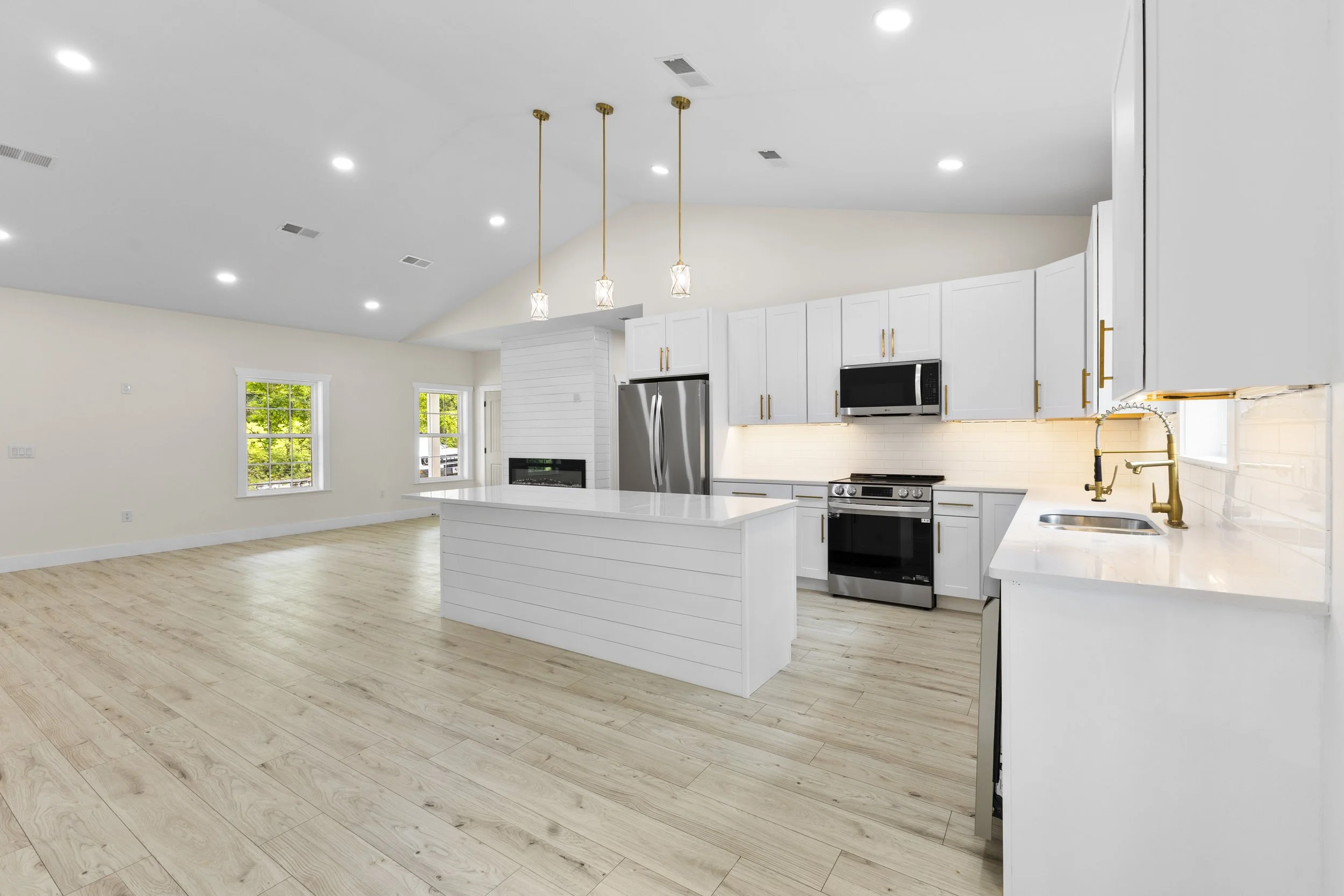 Modern kitchen with white cabinets, black appliances, light wood flooring, and gold fixtures, open to a living area with three windows and white walls.
